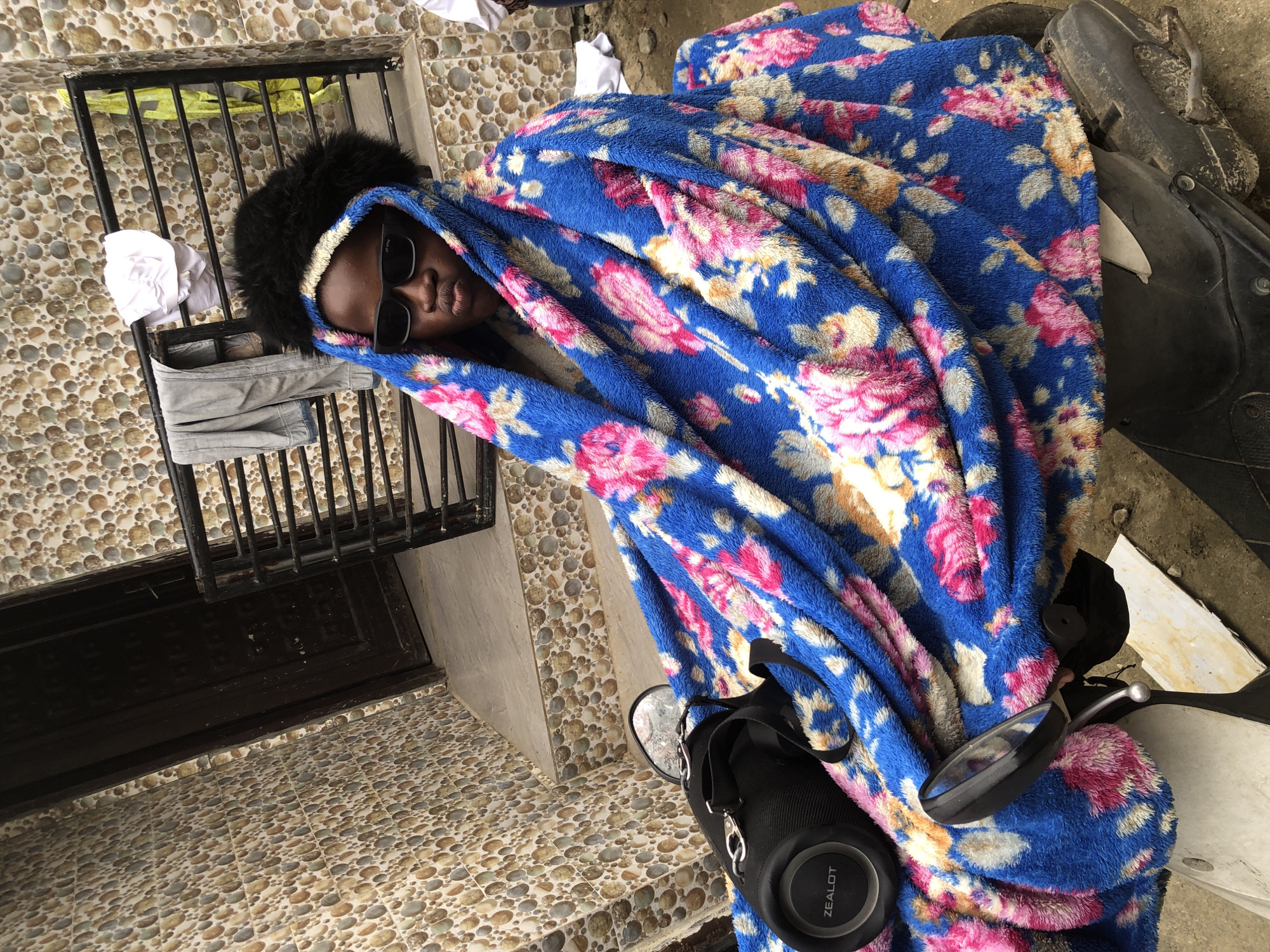 A child, wearing sunglasses and a fluffy black headpiece, sits on a motorcycle covered in a blue blanket with a floral pattern. The child is draped in the blanket, with only their face and hands visible. A black speaker with the brand name "ZEALOT" is attached to the motorcycle. The background features a wall with a mosaic-like pattern of circular tiles, a dark wooden door, and a black metal railing. Clothes are hung on the railing, including a white garment and a grey garment. The scene appears to be outdoors, with a rough, dirty ground surface. The setting is a residential area in Ado, Nigeria.