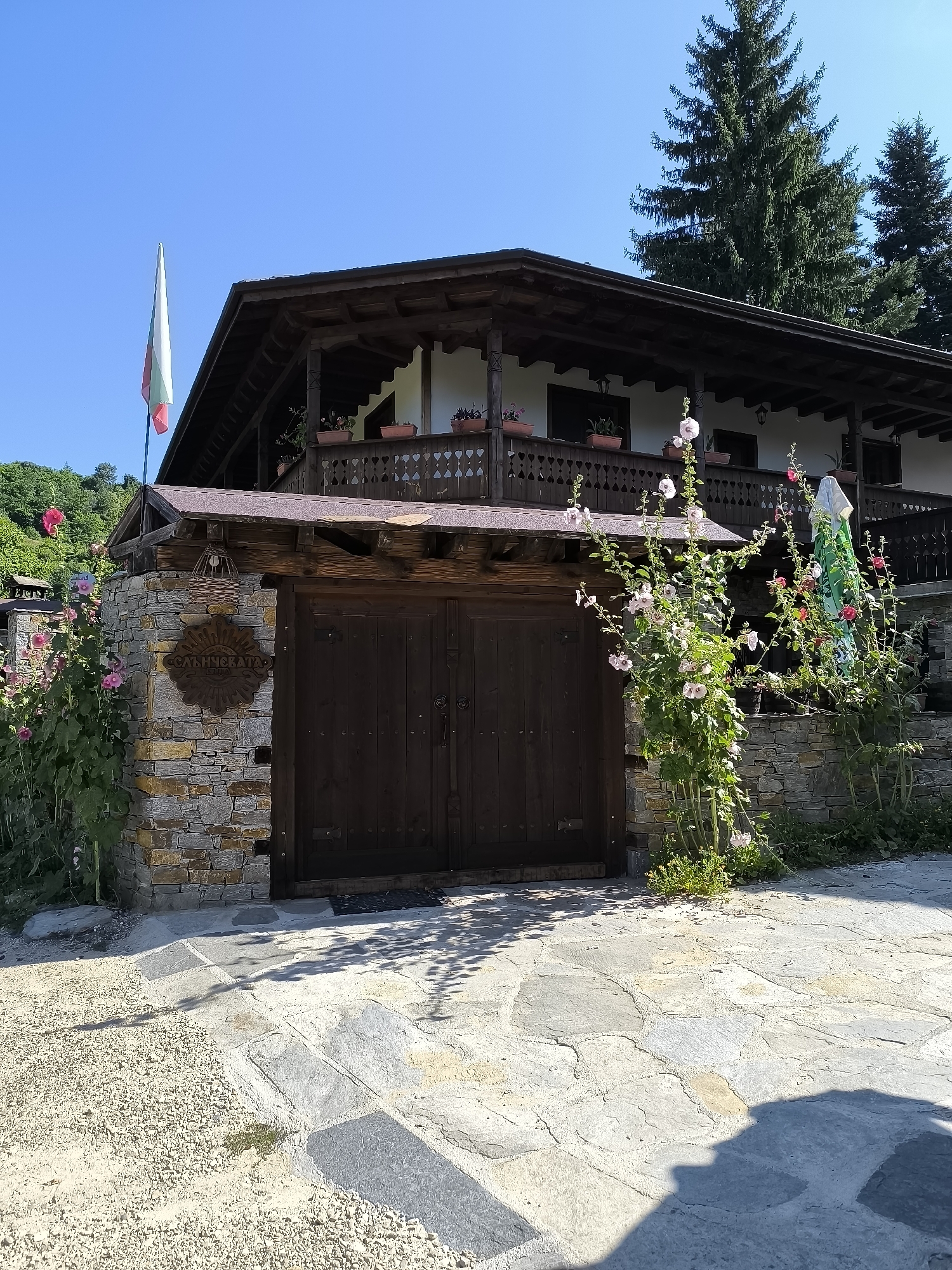 The image shows the facade of a traditional Bulgarian house in the village of Kovachevitsa. The house features a wooden balcony with ornate railings and potted plants. A Bulgarian flag flies from a pole to the left of the house. In front of the house is a stone wall with a large wooden gate. A sign with Cyrillic lettering is mounted on the wall. Tall hollyhock flowers bloom in front of the gate and to the right of the house. The ground is paved with large, irregular flagstones. The scene is set under a clear blue sky, with evergreen trees visible in the background. The overall impression is one of rustic charm and traditional architecture.