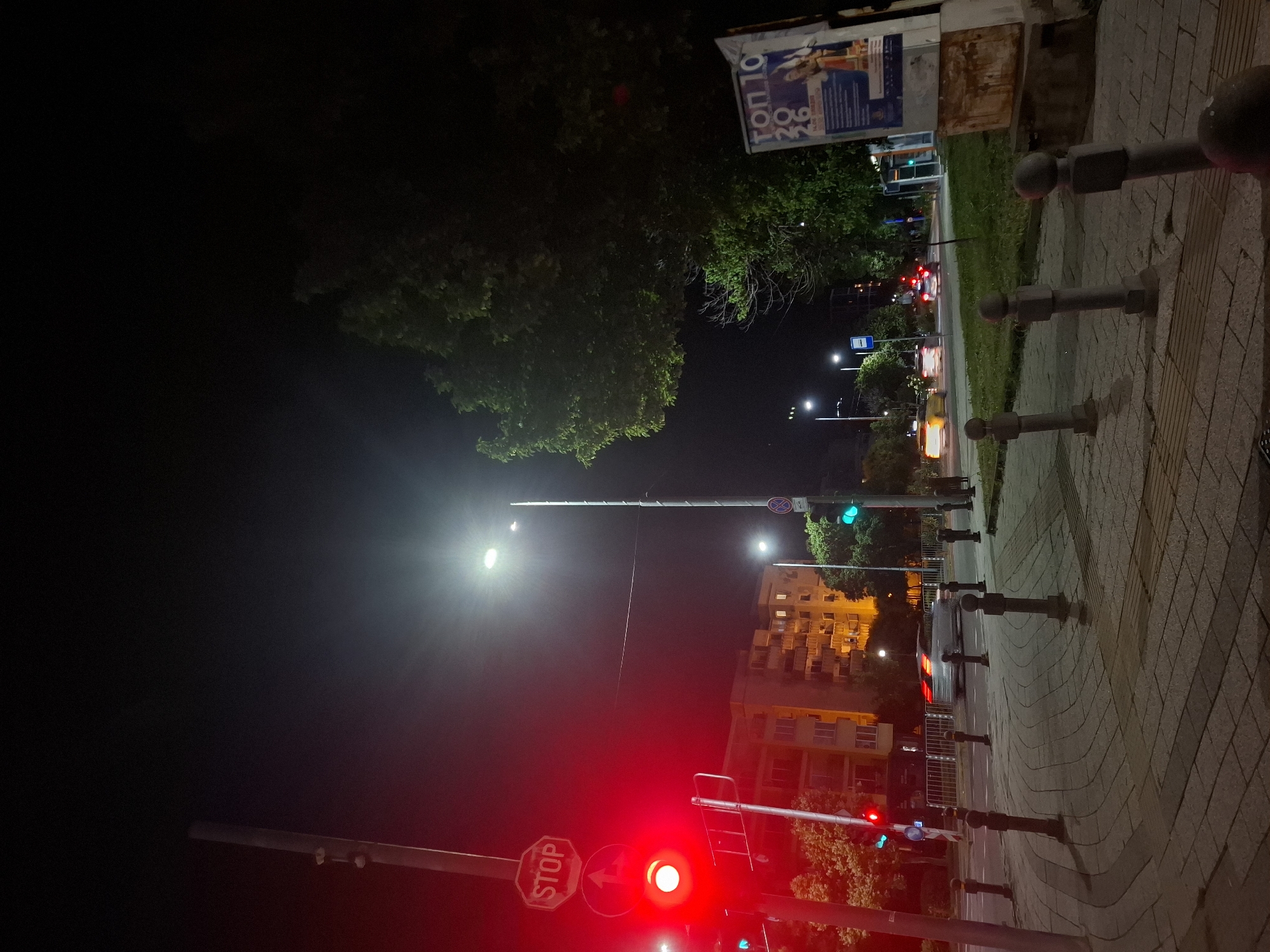 This is a night-time, eye-level, full shot of a city street intersection. A traffic light on the left is illuminated red. A stop sign is positioned above the traffic light. A multi-story apartment building with illuminated windows is visible in the background. Trees line the street and sidewalk. Several streetlights are on. A car is driving through the intersection. A poster with Cyrillic text is attached to a structure on the right side of the image. The foreground consists of a paved sidewalk with bollards.
