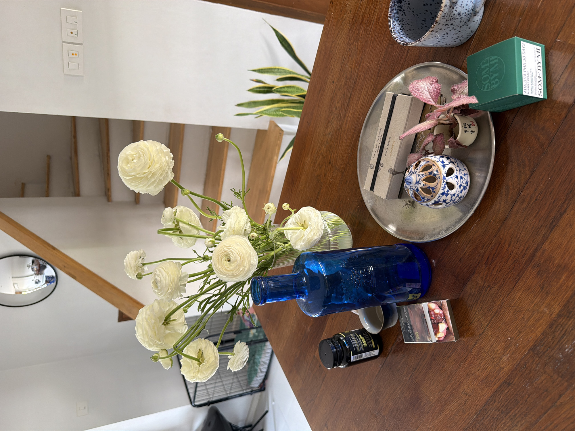 A high-angle, slightly rotated shot features a wooden table surface covered with an assortment of objects. A vibrant bouquet of white ranunculus flowers is arranged in a blue glass bottle vase. Positioned near the vase are small containers or bottles, one being black with a yellow label, and a pack of cigarettes. A silver tray sits on the table, holding a box of incense sticks labeled "TORRENTO WOODY & ANDER", a small plant with reddish-pink leaves in a tiny decorative pot, a decorative ceramic ornament with holes, and a green box labeled "SOME BY MI". A speckled blue ceramic mug is situated near the tray.
The backdrop reveals a white wall with light switches and a wooden staircase. A section of a snake plant is also visible. A black, round mirror is mounted on the wall, reflecting a portion of the interior.
Given the OCR data indicating "INCENSE STICKS" and possible origins like "TONKA BEAN, OUD, MYRAN," coupled with the presence of a "SOME BY MI" product, the objects suggest a space likely furnished with items from or related to beauty, wellness, and potentially imported or niche lifestyle products. There is no indication that the picture was taken in Chang Phueak, Thailand.