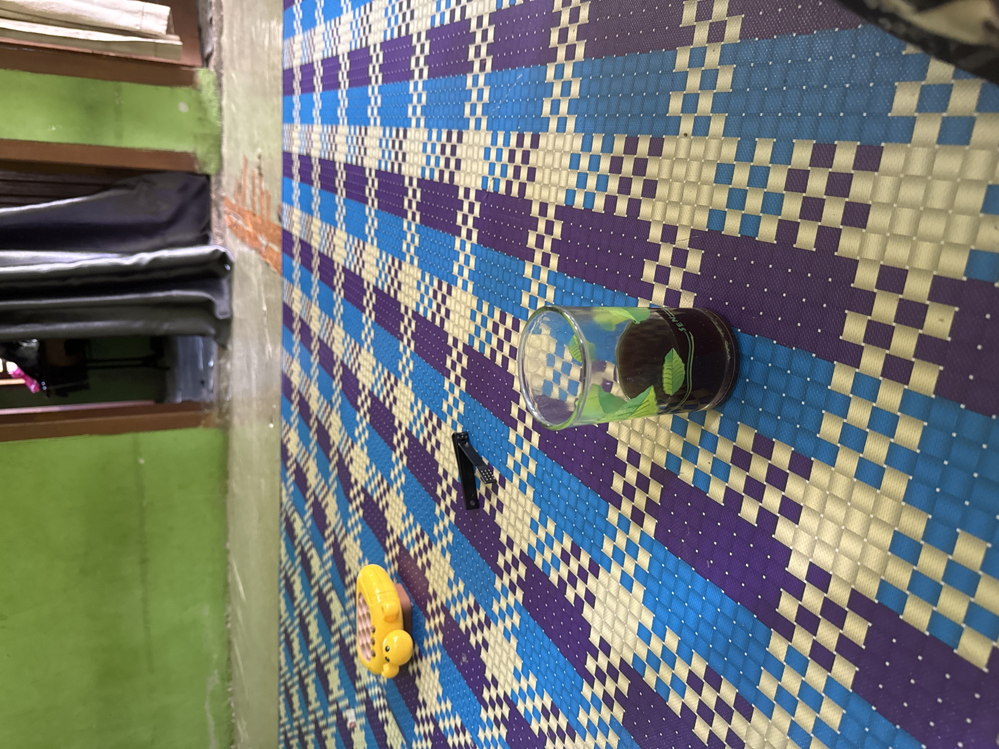 In Babahjurong, Indonesia, an indoor scene is depicted with a focus on a woven mat on the floor. The mat features a repeating geometric pattern of blue, purple, and off-white squares. On the mat, a small yellow toy, possibly a duck, and a pair of black nail clippers are visible. Towards the foreground, a clear glass contains a dark liquid, possibly coffee or tea, and a green leaf decoration is visible inside the glass. The background shows a green wall and a doorway leading to another room, with curtains drawn. The lighting suggests it is daytime.