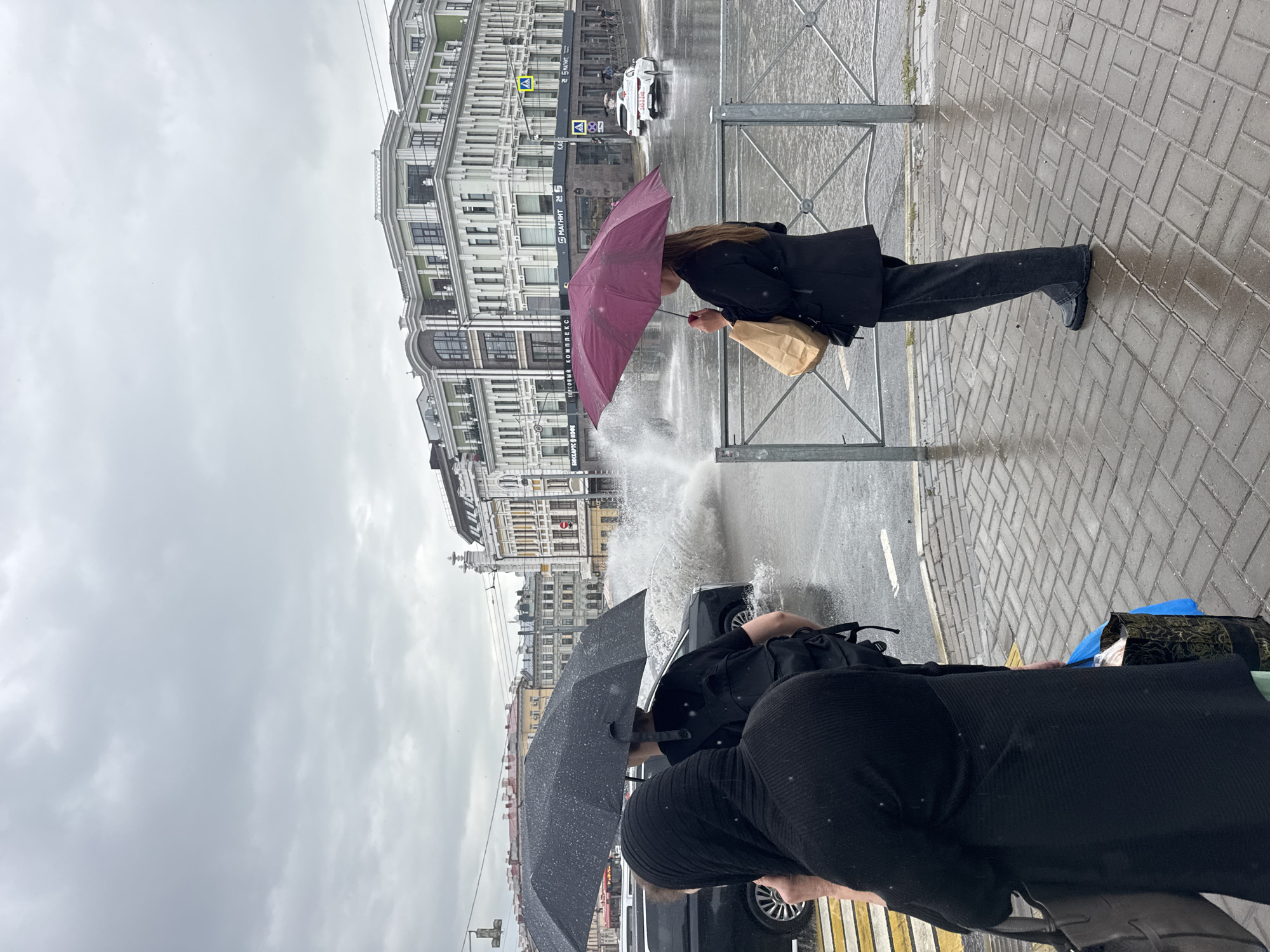 In Kazan, Russia, on a cloudy day, a street scene unfolds with a large puddle on the road, likely caused by a burst water main or heavy rain. A black car is seen splashing through the water. Two people are visible in the foreground, one wearing a black head covering and the other holding a dark umbrella. Further back, a person stands holding a burgundy umbrella and a paper bag, looking towards the water splash. Buildings line the street in the background, with a yellow pedestrian crossing visible. A white car is seen driving in the distance. The ground is paved with bricks, and a metal fence separates the sidewalk from the road.