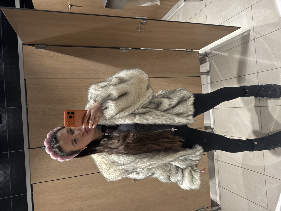The image shows a female individual taking a mirror selfie in what appears to be a public restroom. The precise location within the city of Akseno-Butyrki, Russia, is not ascertainable from the visual information provided.
The subject is a woman with long, dark brown hair, styled with a pink, segmented headband. She is wearing a voluminous, light-colored faux fur coat, appearing in shades of white, grey, and black. Underneath the coat, a dark-colored, zippered top or hoodie is visible. She wears black leggings and black, lace-up boots with prominent soles. The woman is holding an orange smartphone in her right hand, angled towards the unseen mirror to capture her reflection. Her lips are pursed, and a light-colored manicure is visible on her fingernails. A silver-toned ring is present on her left ring finger.
The environment consists of light-colored wooden or laminate partitions for restroom stalls. The floor is covered with large, light-colored square tiles. Above the stall partitions, the wall features dark-colored, possibly black, tiles or panels. On the right side of the image, a metal toilet paper dispenser and a white paper towel dispenser are mounted on a stall wall. Two stall doors are visible, one partially open, with silver-toned door handles/locks and floor-mounted pivots or hinges.