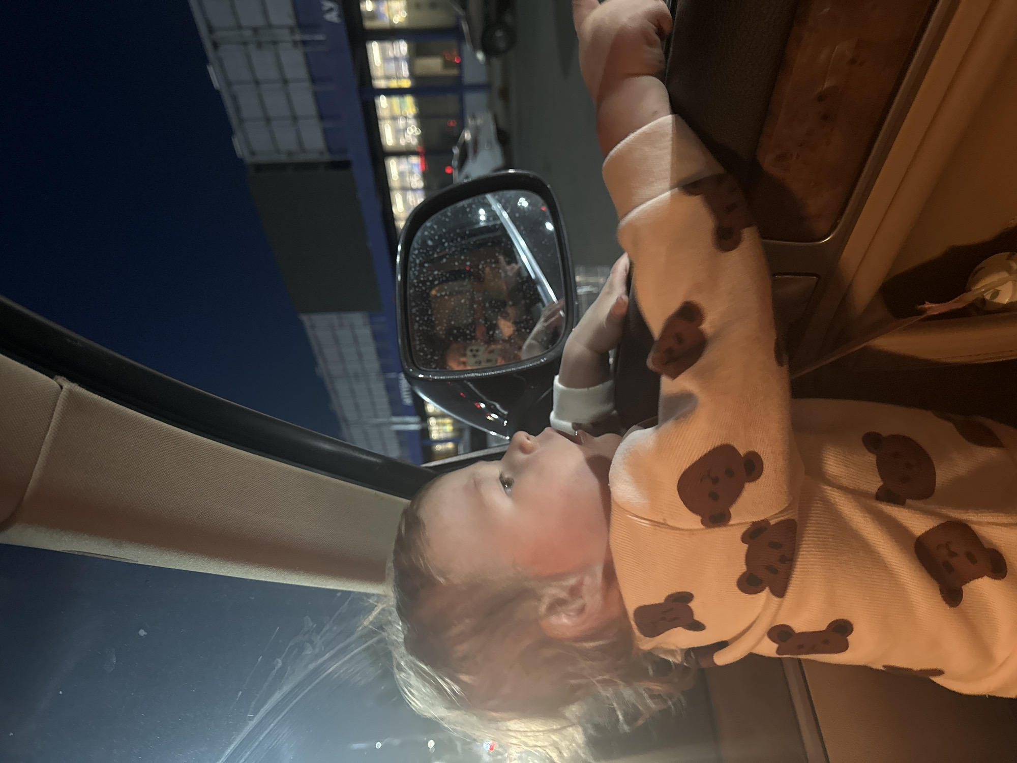 A baby in a teddy bear-print onesie is in a car, looking toward the side mirror. The reflection in the side mirror shows other passengers and the exterior environment. Outside of the car, there are buildings, street lights, and street signs. A sign appears to read "AY.." This suggests the location of the photo is Nizhny Novgorod, Russia. The sky is dark.