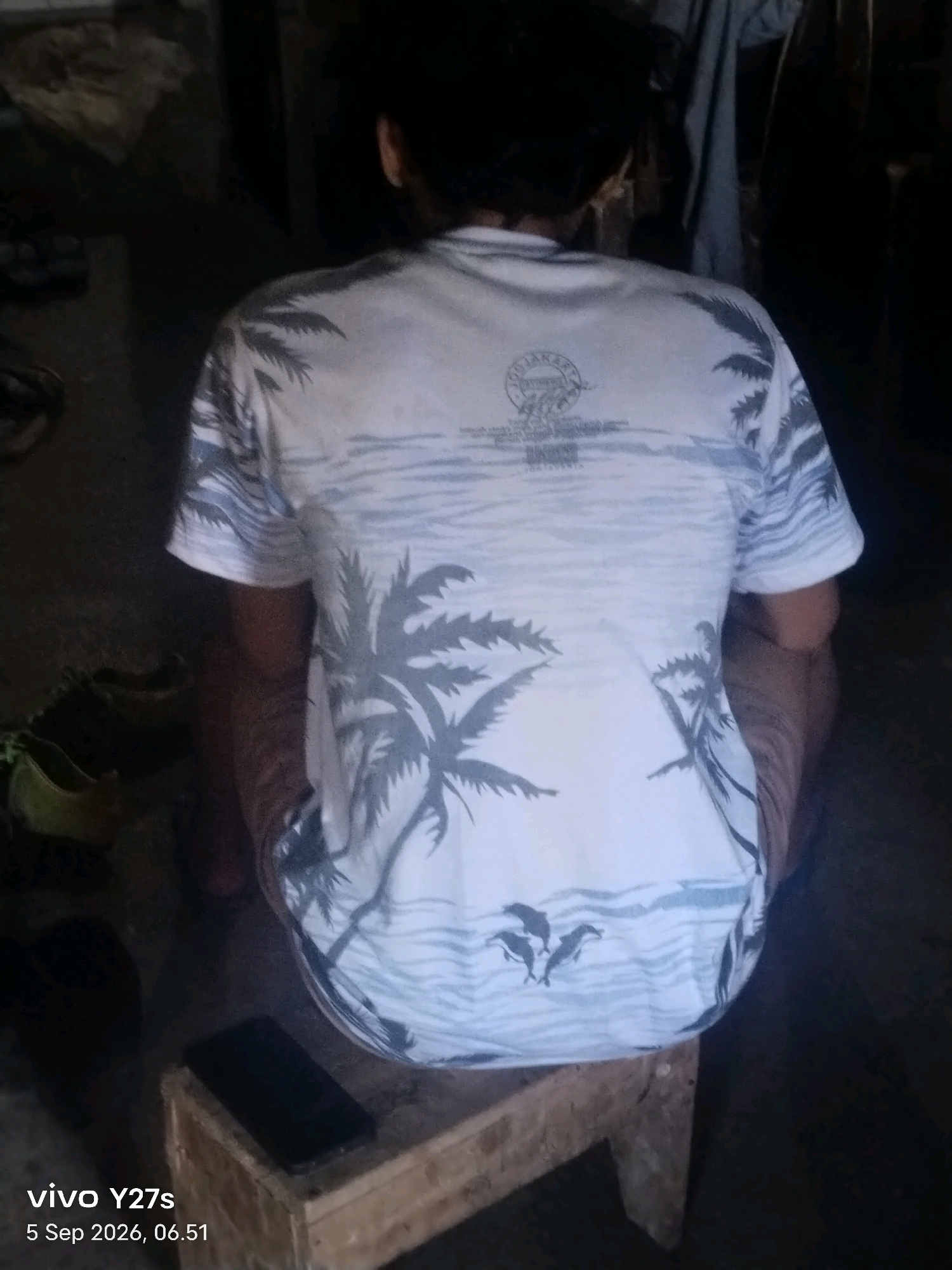 A person, viewed from behind, is seated on a wooden stool in a dimly lit indoor environment. The person is wearing a white t-shirt with a tropical pattern featuring palm trees, waves, and dolphins. A dark-colored smartphone rests on the surface of the stool. The background appears to be cluttered with various items, including what might be fabric hanging and some indistinct objects on the floor. The timestamp overlay indicates the image was taken in Tamansari, Indonesia on September 5, 2026, at 06:51.
