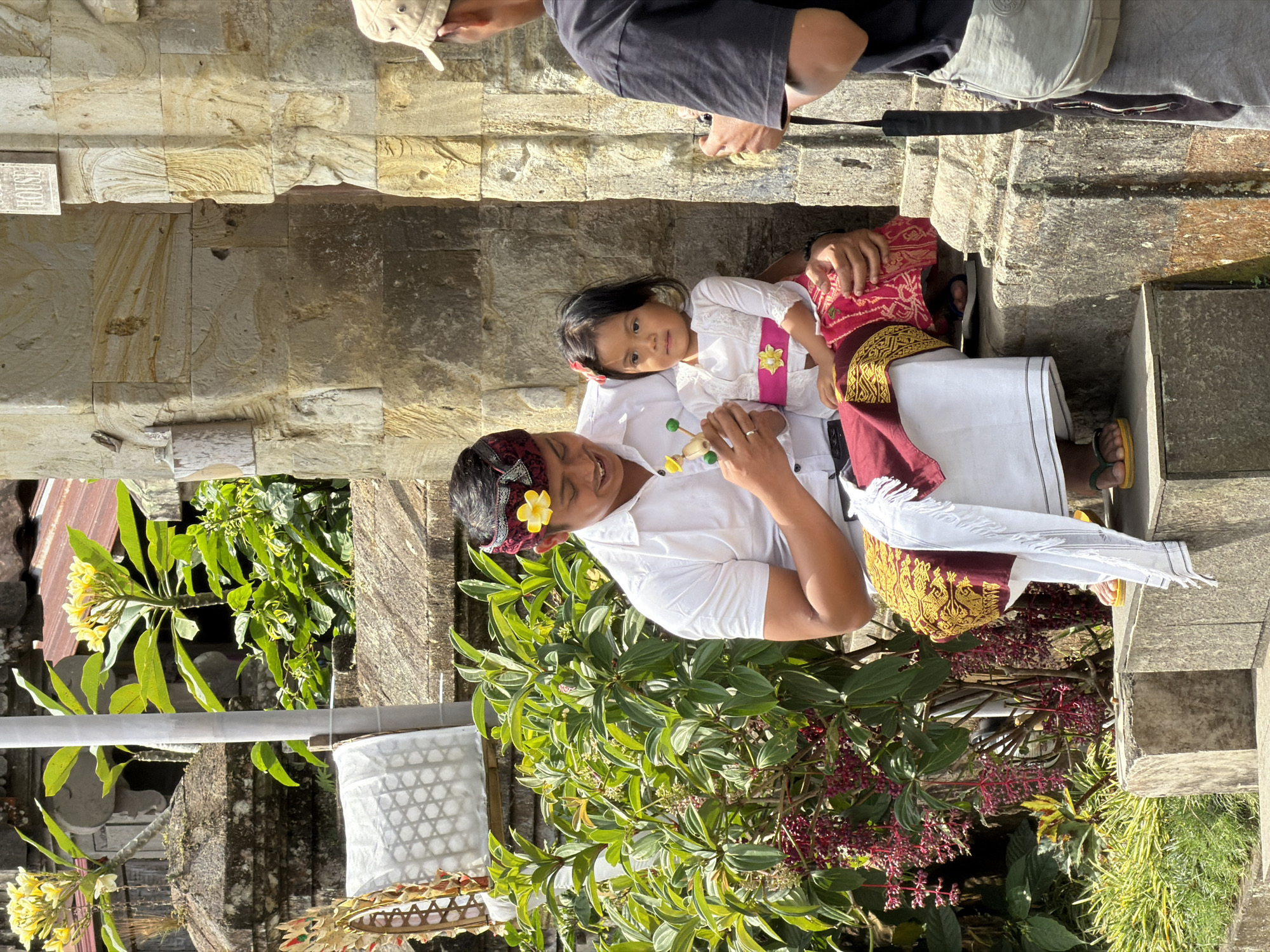 In Susut, Indonesia, a man wearing traditional Balinese attire, including a patterned headcloth with a yellow flower and a white shirt, sits on stone steps. He holds a small, wooden toy in his hands and appears to be interacting with a young girl seated on his lap. The girl is dressed in a white top with a pink sash and a matching pink skirt. A man wearing a dark t-shirt and a beige cap stands to the right, holding a camera and seemingly photographing the pair. Behind them, a stone structure with intricate carvings is visible, along with lush green foliage and blooming yellow flowers. The scene is set outdoors, in what appears to be a temple or historical site, with the sunlight casting shadows on the stone surfaces.