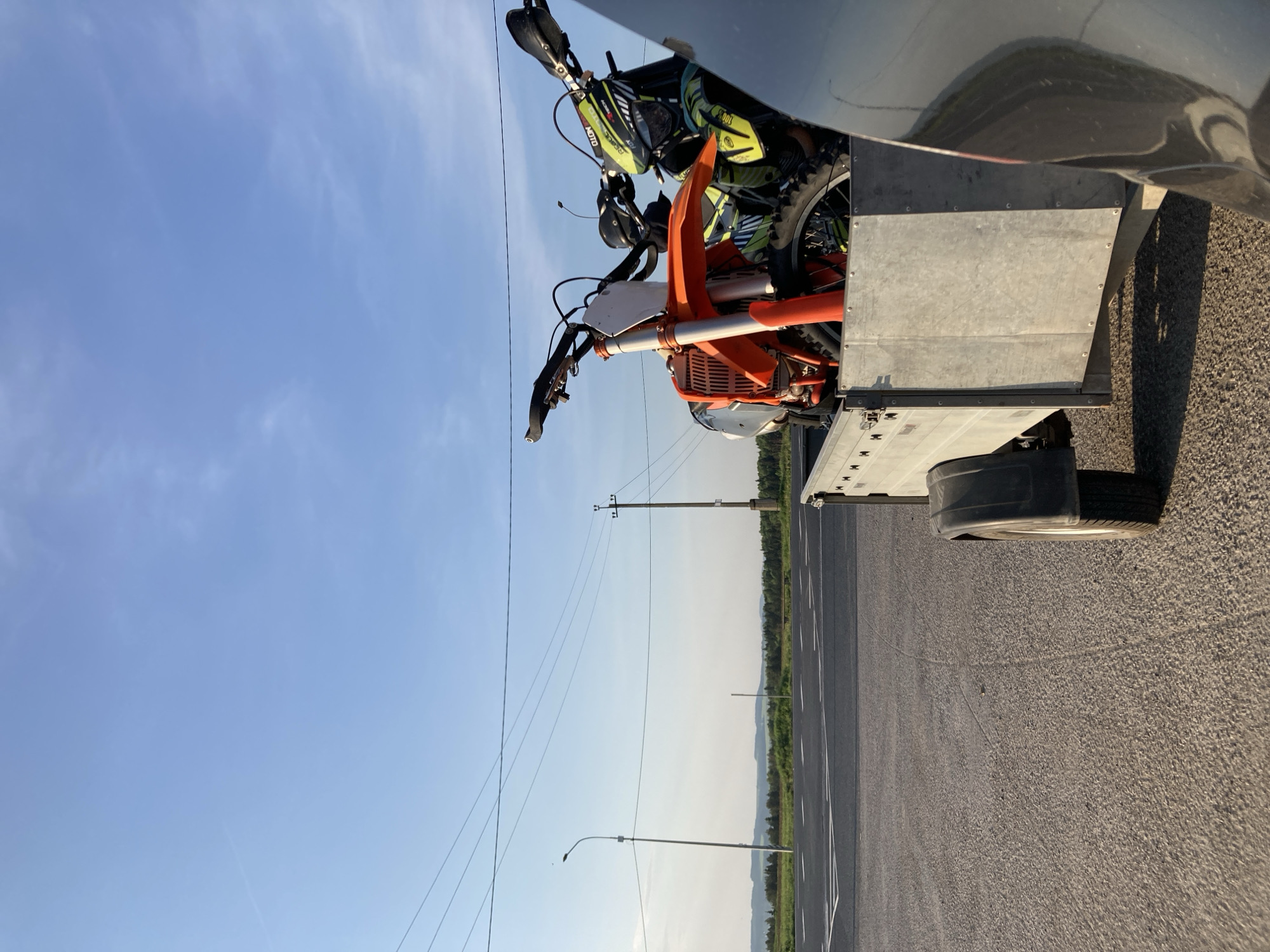 The image shows two motorcycles on a trailer, parked on an asphalt road. The road appears to be a highway or major thoroughfare, with road markings visible. Power lines and poles run alongside the road. In the background, there's a glimpse of vegetation, possibly trees and hills, suggesting a rural or semi-rural setting. The sky is blue with some wisps of clouds. The location is Verkh-Sosnova, Russia.