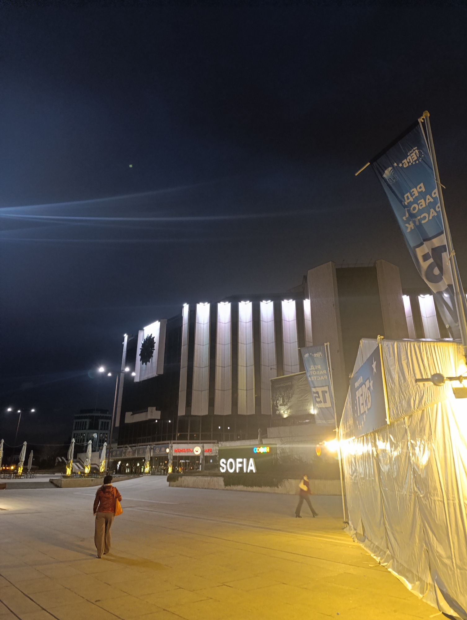 The image depicts an outdoor scene at night in Sofia, Bulgaria. In the background is the National Palace of Culture (NDK), illuminated with vertical lights and a large emblem. "SOFIA" is written in large, illuminated letters near the building. In the foreground, a person is walking away from the camera. Another person is walking in the background, past what appears to be a cafe. The ground is paved, and a white tent structure with advertising banners is on the right side of the image, brightly lit. A blue flag with reversed writing is also prominent on the right. The sky is dark, with some light flares.