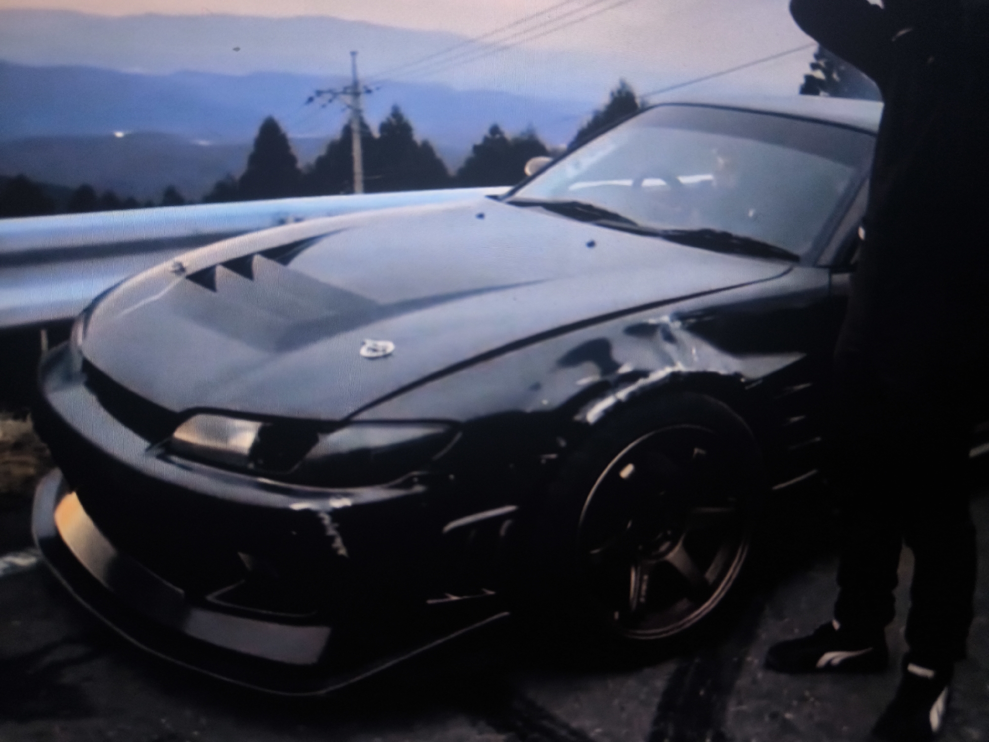 A black sports car is parked on a paved surface, possibly a road or track, with tire marks visible on the ground. The car has a modified hood with vents and racing-style hood latches. A person, dressed in dark clothing and black athletic shoes with a white stripe, stands next to the car, with their arm raised. The background shows a landscape with mountains, trees, and a utility pole with wires. The lighting suggests it is either dusk or dawn. The specific location is unknown.