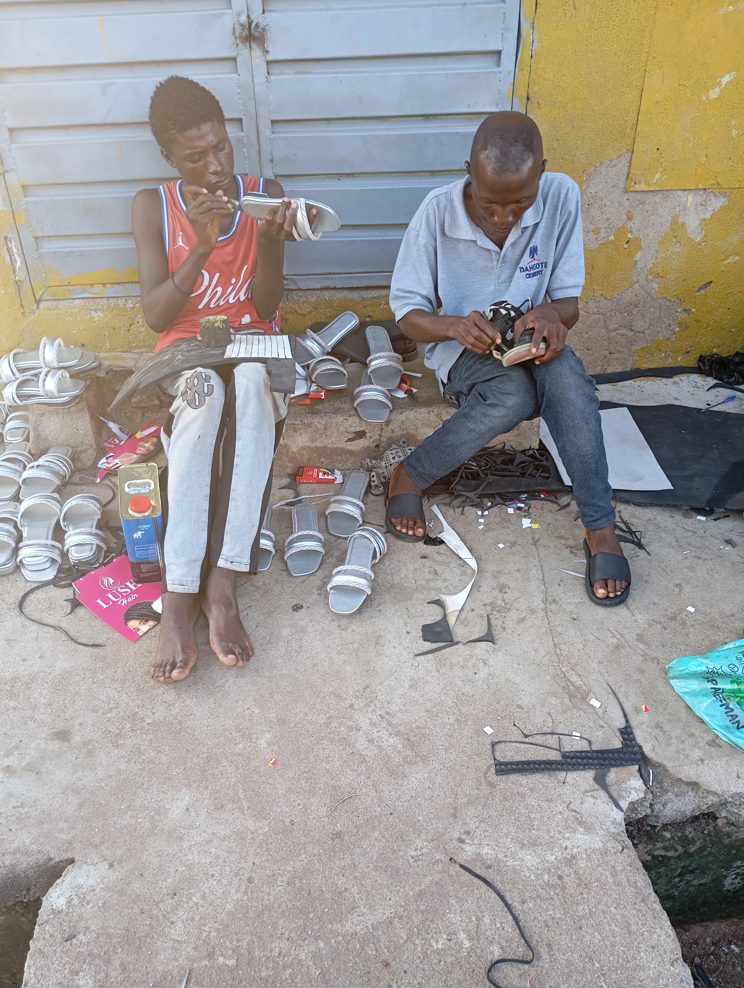 Two individuals are observed sitting on a concrete surface in Shendam, Nigeria, engaged in the process of crafting sandals. The individual on the left, wearing a red basketball jersey and light-colored pants, is intently working on a sandal, holding it in their hands. A collection of finished sandals is arranged to their left. To their right, a can of yellow liquid and a pink booklet with an image of a woman are placed on the ground. The individual on the right, dressed in a gray polo shirt and dark trousers, is also working on a sandal, holding it with both hands. Numerous sandals are spread around them on the ground, along with pieces of leather and tools. The environment appears to be an outdoor setting, possibly a street or market stall, with a textured yellow wall in the background and a corrugated metal structure above. The ground is concrete, with some debris and scattered materials related to sandal making.