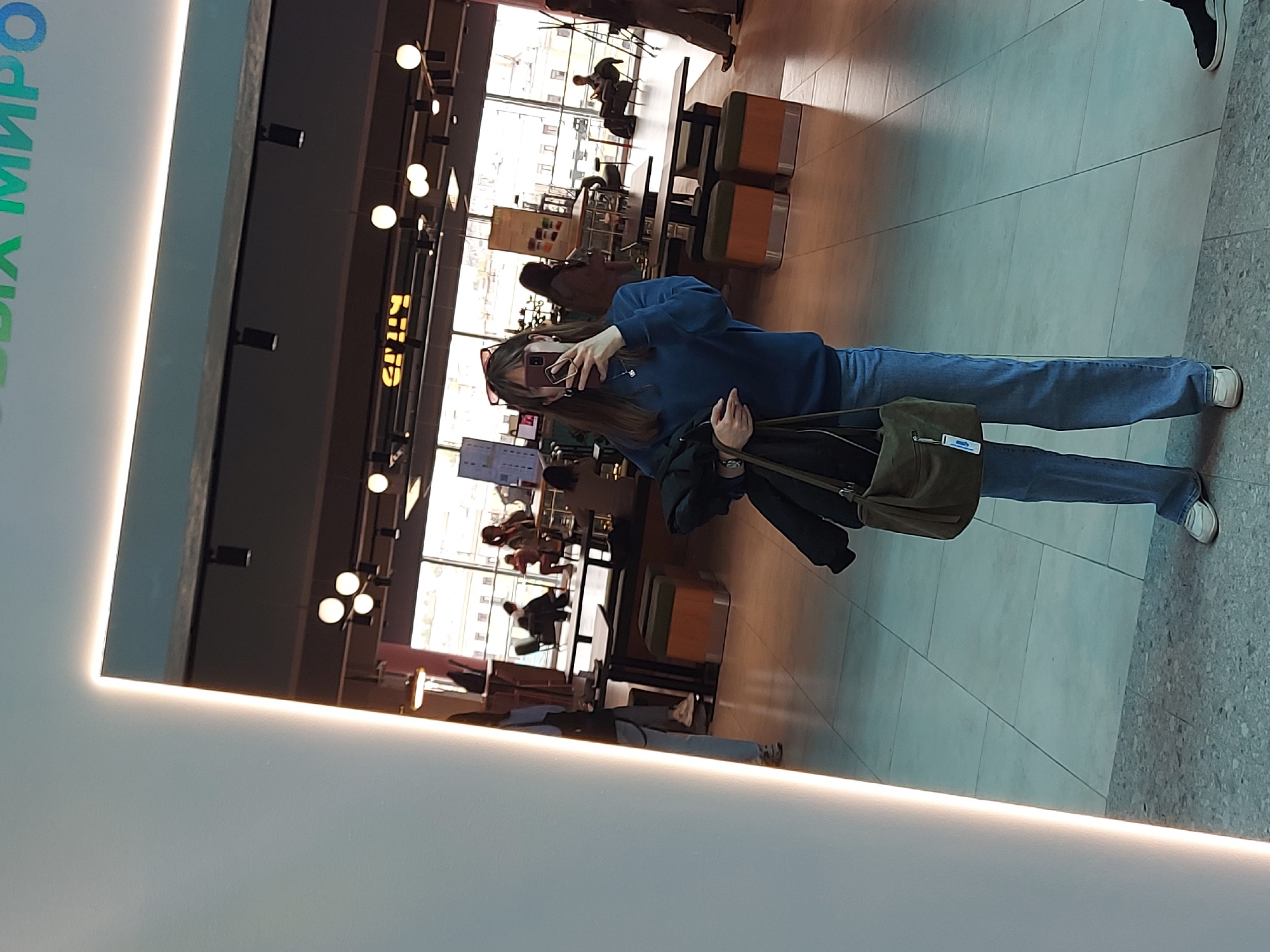 A person in a blue sweater and jeans is taking a photo of themself in a mirror. They are holding a phone in their right hand and a dark jacket and green bag in their left hand. The mirror is surrounded by a bright, white frame.
The reflection shows a brightly lit interior with a window and tables and chairs. People are sitting and standing in the background. Overhead lights are visible.
Based on the sign visible and the architectural style, this appears to be located in Saint Petersburg, Russia.