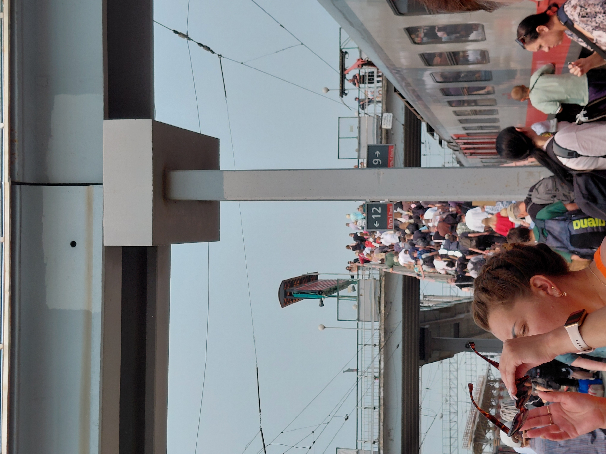 A high-angle, medium shot captures a bustling scene at a train station in Novosibirsk, Russia, with a prominent overhead pedestrian bridge and a train on the right. In the foreground, a woman with brown hair, wearing an orange top and a smartwatch, holds a pair of tortoise-shell sunglasses to her face, her left hand displaying rings. Behind her, a crowd of people ascend a staircase to the left, some carrying luggage. Signs indicating "← 12" and "9 →" are visible on a pillar. A silver train with red accents, its windows reflecting the sky, is parked to the right of the frame. The environment includes overhead wires and the grey structure of the elevated walkway and station. The sky is overcast and pale.
