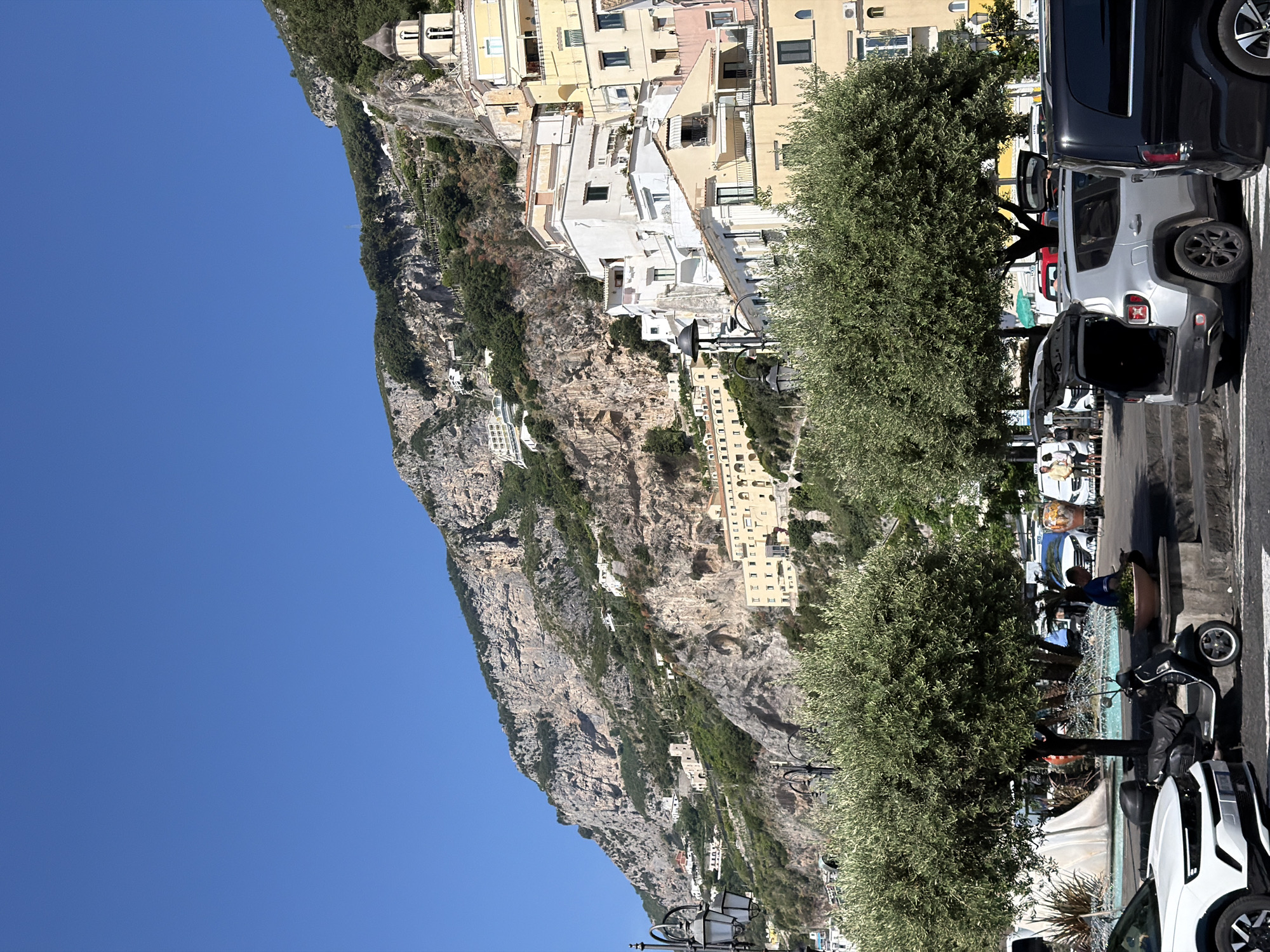 The image depicts a street scene in Pogerola, Italy, set against a backdrop of a steep, rocky cliff face dotted with buildings. The sky above is clear and blue.
A row of parked cars lines the right side of the street, including a dark van and a white SUV with its rear door open. Two large, bushy trees stand between the cars and the cliff face, partially obscuring the view of the buildings.
On the street, a partially visible water feature or fountain stands next to a parked scooter. People are walking around, suggesting it is an active public space. The buildings visible on the cliff are a mix of white and yellow structures.