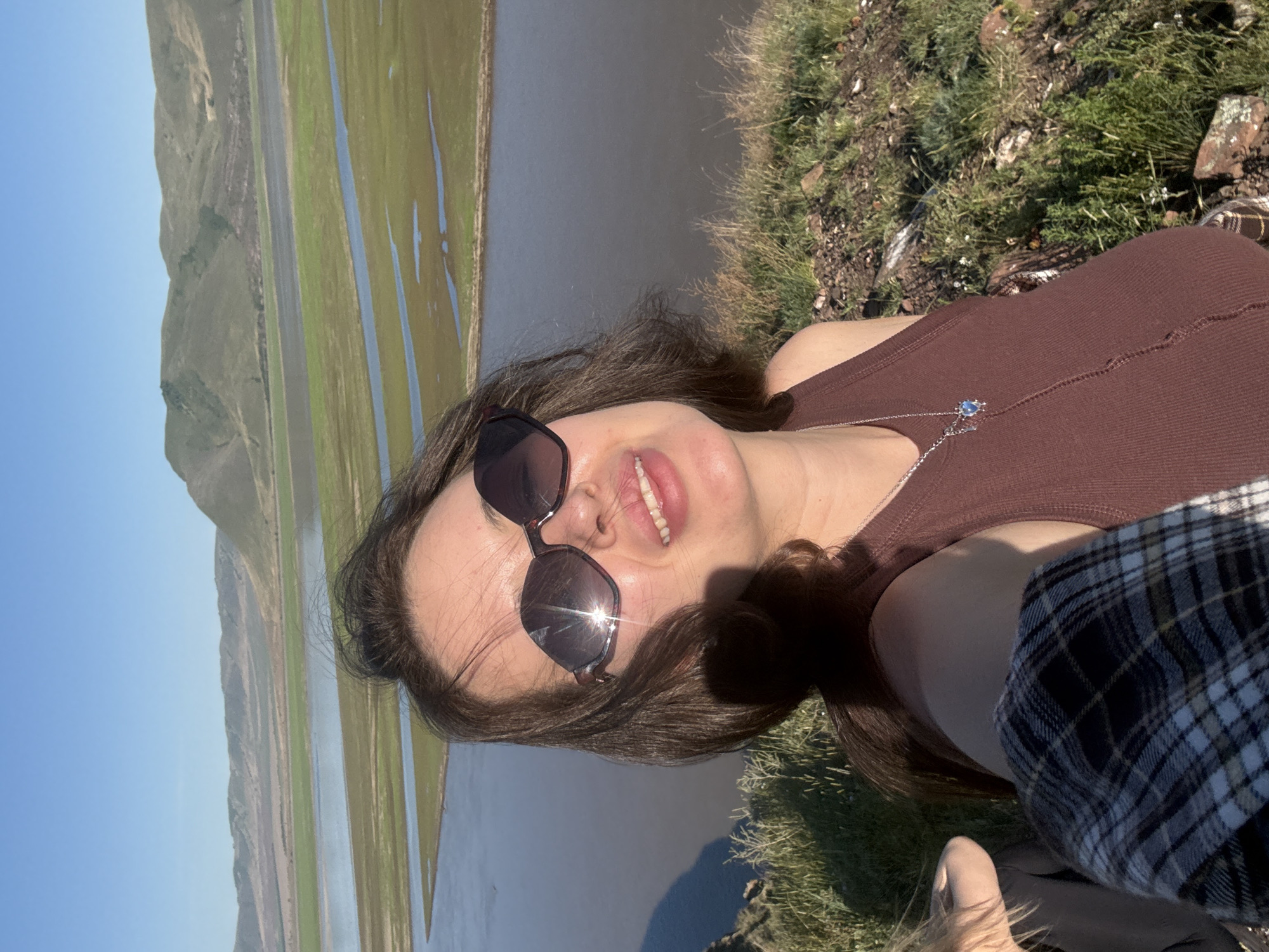 The image is a bright, sunlit selfie featuring a young woman smiling broadly outdoors. The photo is rotated, appearing on its side, with the woman positioned on the right and the landscape to her left.
The main subject is a woman with long, dark hair, wearing a brown ribbed tank top and dark sunglasses. A silver necklace with a small blue pendant is visible around her neck. Sun glare reflects prominently off her sunglasses, indicating a bright sunny day. Her wide smile conveys a sense of happiness and enjoyment. Part of a plaid shirt, possibly black and white or dark blue and white, is visible in the bottom right corner, suggesting it might be worn by another person next to her or draped over her.
The background reveals a vast, open landscape under a clear blue sky. The woman appears to be situated on a grassy, rocky elevated area, overlooking a wide body of water—possibly a river or lake—with lush green plains and fields stretching beyond it. Distant, undulating hills or mountains line the horizon. The sunlight casts subtle shadows, suggesting a bright day.
The setting is a scenic natural environment, consistent with the provided location context of Ust'-Abakan, Russia. The overall impression is one of leisure and appreciation for nature on a beautiful, clear day. There is no visible text in the image.