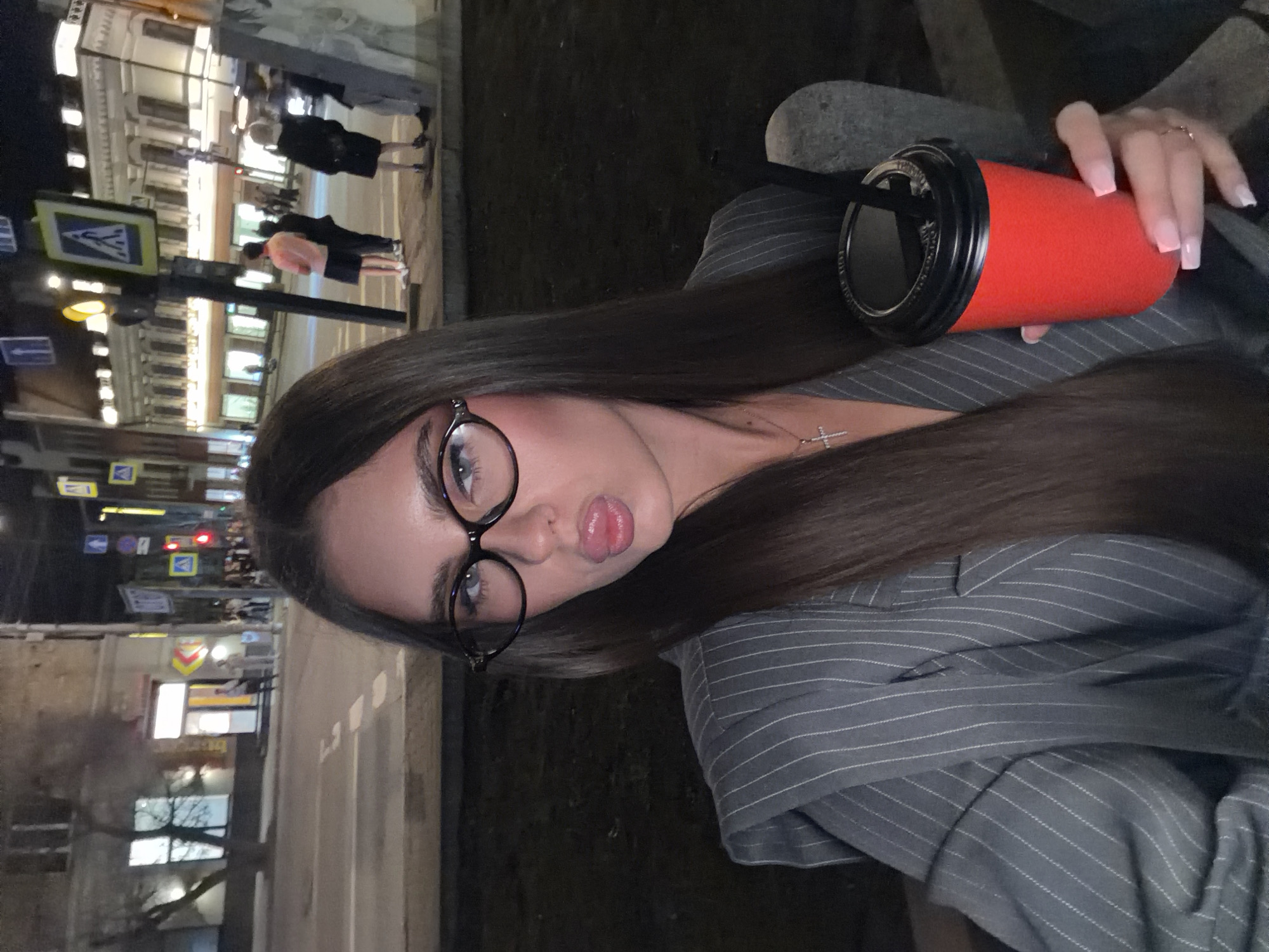 A young woman is holding a red cup with a black lid and straw. She is wearing glasses and a striped gray blazer. The photo appears to be taken at night in Krasnoyarsk, Russia, based on the city's street signs and architecture in the background. Pedestrians and street lighting are also visible.
