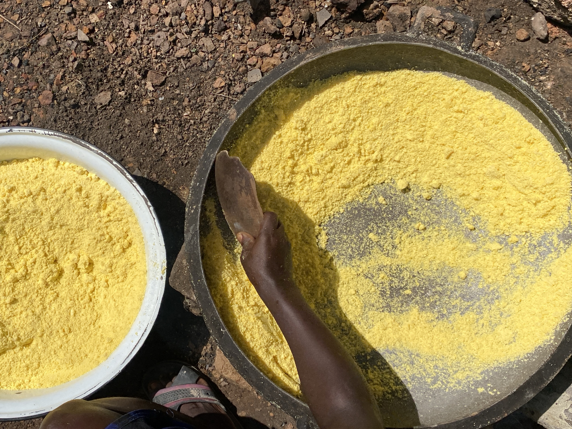 From a high angle, the image shows a person working with a large bowl and a shallow pan filled with yellow powder, possibly flour or cornmeal. The person is using a flat, wooden tool to manipulate the powder within the dark-colored pan. To the left is a white bowl nearly filled with the same yellow powder. The scene takes place outdoors on bare ground covered with small rocks. A foot wearing a sandal is also visible at the bottom of the frame. The location is presumed to be Ufu Esioni, Nigeria. No specific actions or interactions are evident other than the individual using the tool to work with the powder. The surrounding environment is barren and suggests a rural or less developed setting.