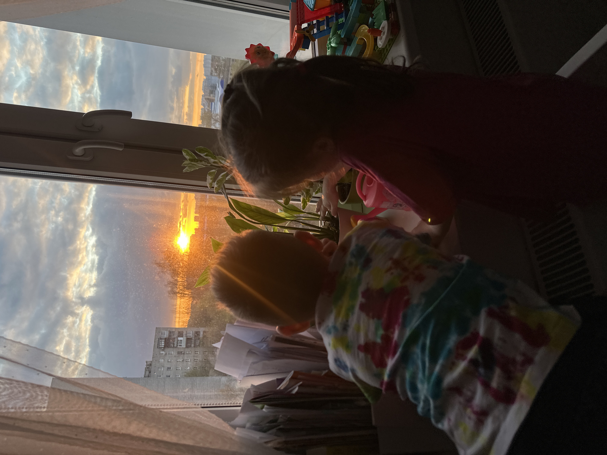 An indoor shot frames two young children at a window during sunset in Tomilino, Russia. The low sun casts a brilliant orange glow across the sky, illuminating clouds and a distant cityscape with a multi-story building. The interior is dimmer, creating a high-contrast scene.
On the left, a sheer curtain or blind hangs, while white window frames and handles are visible. A houseplant sits on the windowsill, near which one child, seen from behind with dark hair and wearing a pink or maroon top, appears to be interacting with the plant or a small pink watering can also on the sill. To their left, another child with lighter hair, wearing a colorful tie-dye shirt, is also by the window, possibly looking out or at a stack of papers and magazines piled on the windowsill. Beyond the children and plant, a vibrant building toy is situated on the right side of the windowsill. A dark heating vent or radiator is partially visible on the far right. The overall atmosphere is one of peaceful domesticity, with the children captivated by the dramatic sunset view. No discernible text is visible in the image.