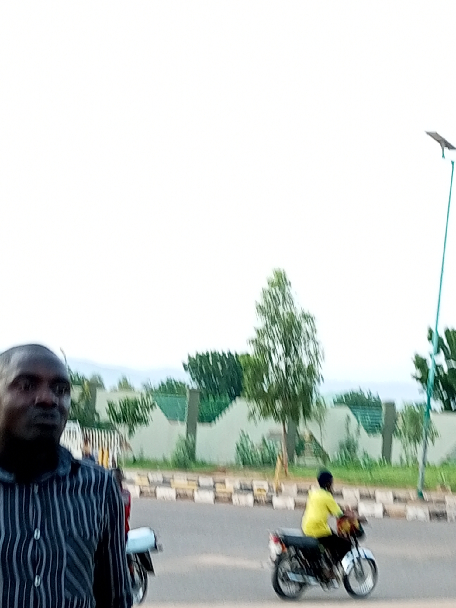 In Gombe, Nigeria, a man in a striped shirt stands in the foreground, looking to his left. Behind him, a person in a yellow shirt rides a motorcycle, heading towards the right of the frame. The motorcycle has a white front fender and white stripes on its wheels. A light blue scooter is parked behind the man in the striped shirt. The background features a street with a curb and a fence line with green foliage. A tall, thin street lamp with a solar panel is visible on the right side of the image. The sky is overcast and light.