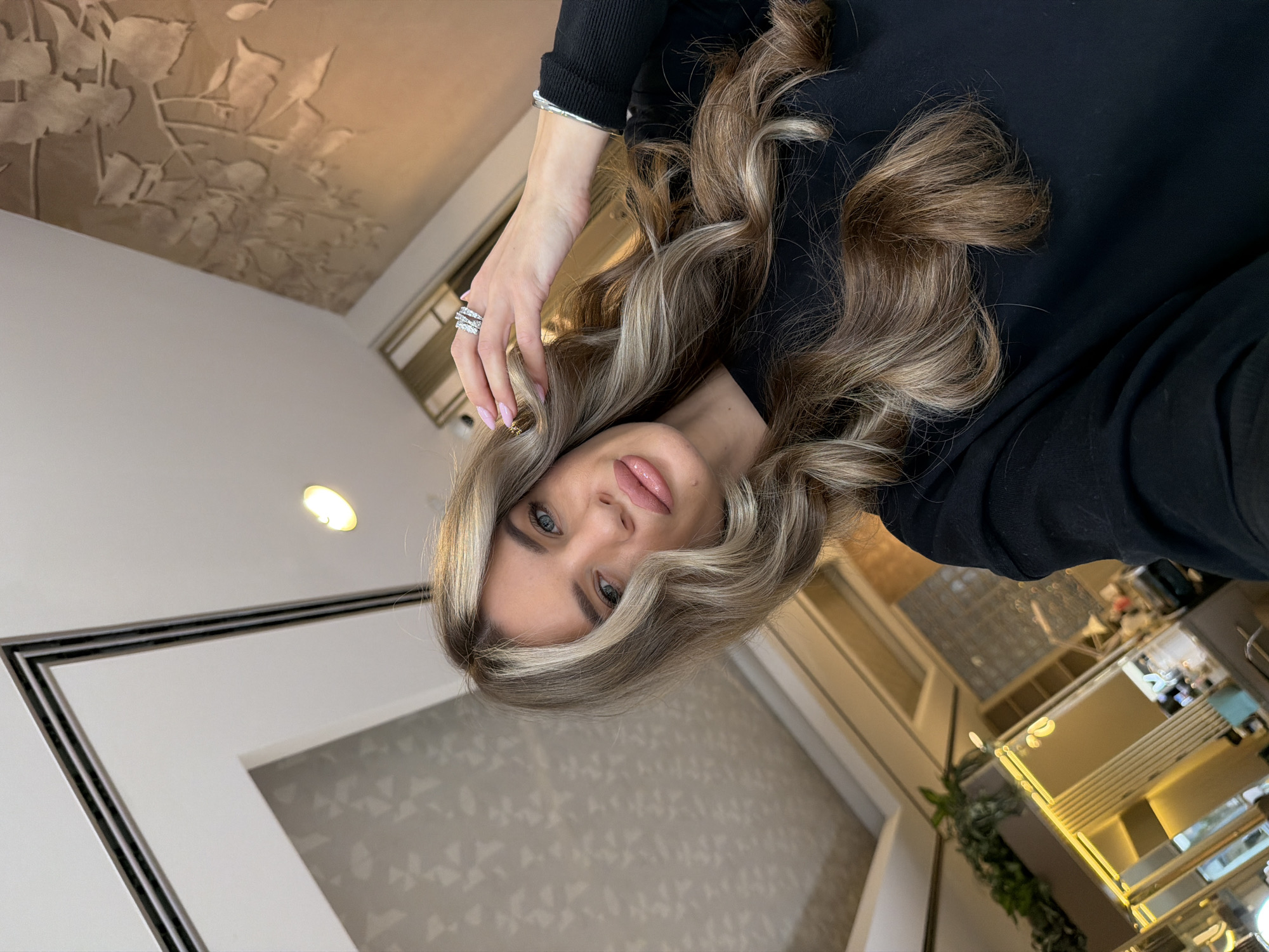 A woman with fair skin, blue eyes, and light brown hair with blonde highlights, wearing a black top and silver jewelry, is the central subject of a photograph taken indoors. The background reveals elements of an interior space, including a patterned wall, a framed artwork, a decorative ceiling, and partial views of furniture. There is no verifiable information in the image to suggest a location in Berlin, Germany.