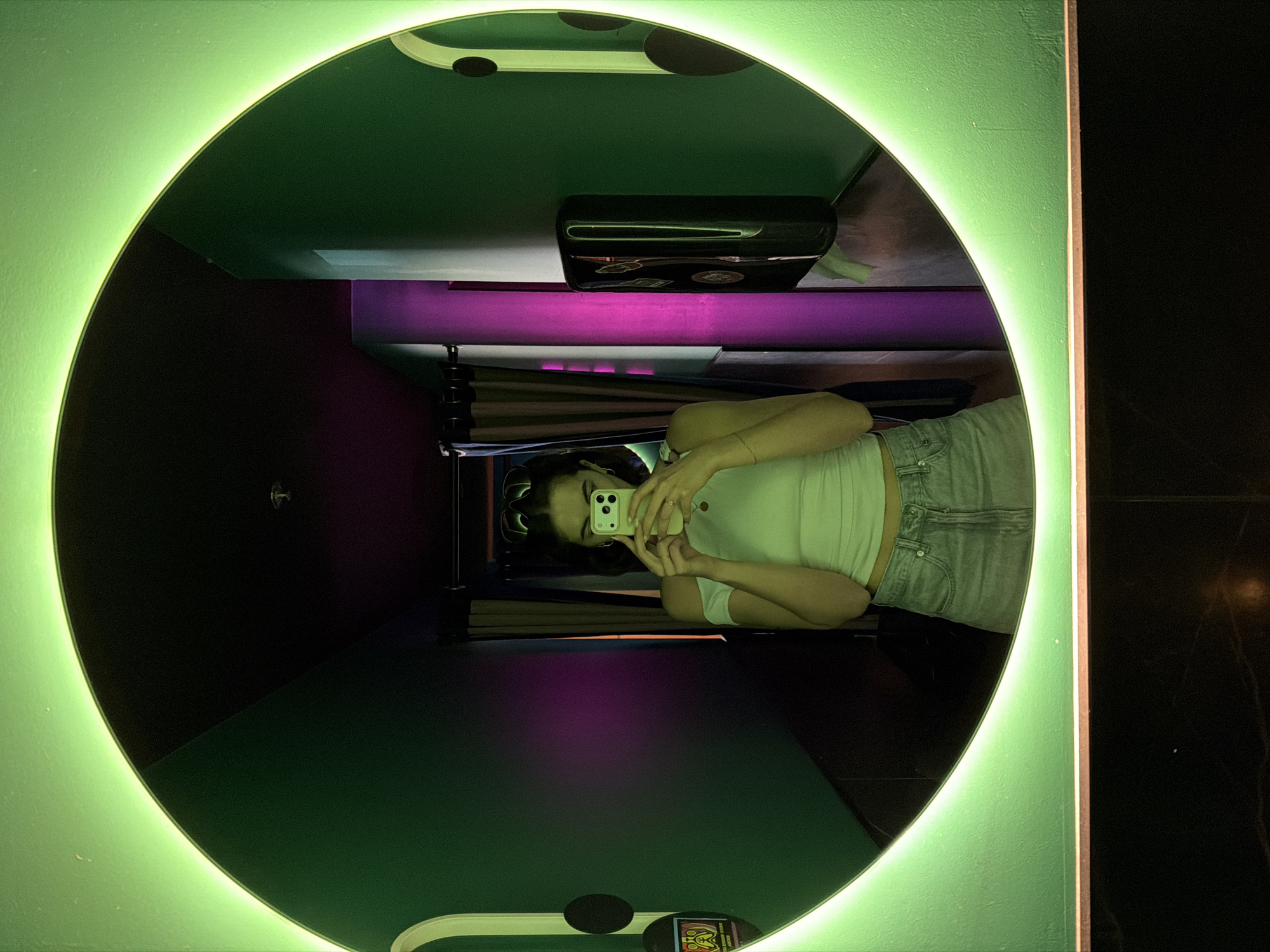 A young woman is taking a selfie in a green-lit oval mirror. The mirror is in a space with dark green walls and a ceiling with black polka dots. There is a shelf above her head with a black case. The lighting alternates between green and purple. She is wearing a white top and light-colored jeans. The room has a black marble-like surface to the right of the mirror. Based on the distinct aesthetic, the location is likely a restroom or photo booth in a trendy bar or club in Montreal, Canada.