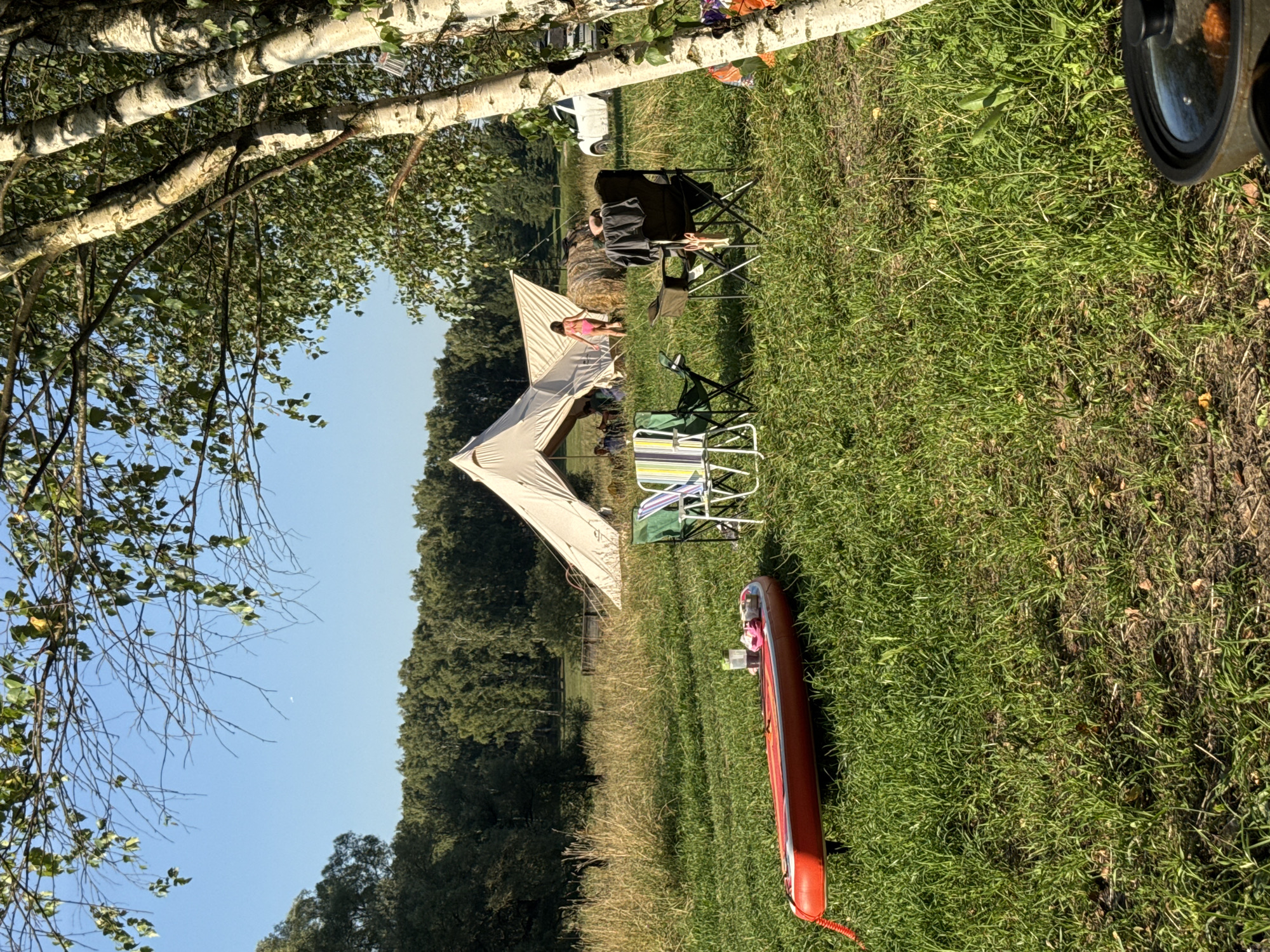 This image depicts an outdoor scene in Kuzemkino, Russia, likely a campsite. In the foreground, a red inflatable paddleboard rests on green grass. Several camping chairs are also present, some unoccupied and others with people seated. A large, off-white bell tent is visible in the middle ground, with figures near its entrance. Behind the tent, a round hay bale is situated, and a white car is parked to the right. A line of trees forms the background, with a clear blue sky above. The overall environment is grassy and appears to be a rural or park-like setting.