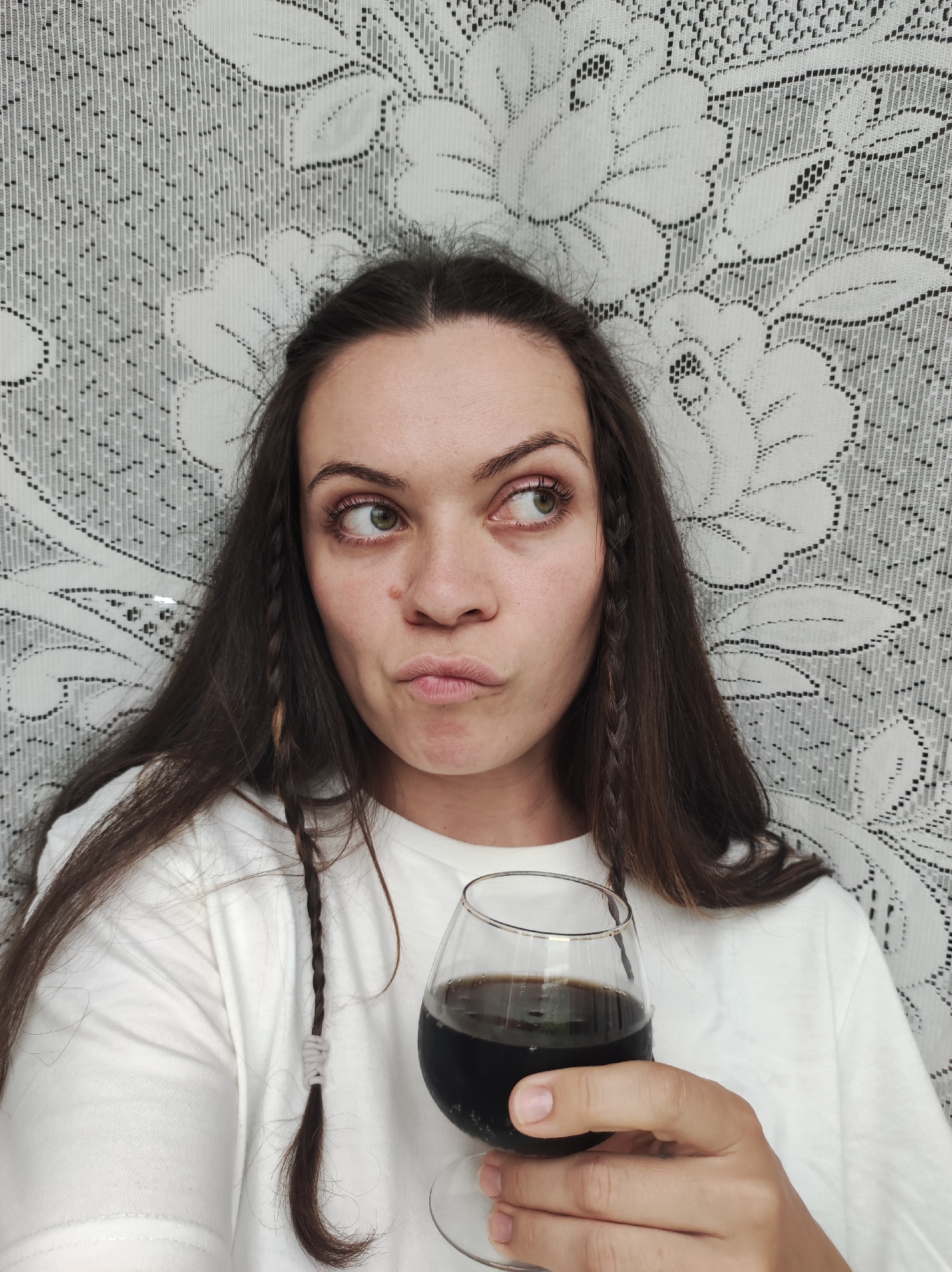 An adult female with long, dark brown hair is positioned from the chest up against a white lace curtain backdrop. She holds a clear stemmed glass containing a dark, opaque liquid, possibly cola or stout, in her right hand. Her light-colored eyes, appearing greenish-hazel, are directed sharply to her right (viewer's left), while her lips are pursed into a prominent pout. Two thin braids, each secured with a light grey or white wrap, are visible among her loose hair, one on either side of her face. A small, light-colored lesion or blemish is present on the bridge of her nose, slightly to the right side. She is wearing a plain white crew-neck t-shirt. The background displays an intricate, repeating floral and geometric lace pattern, characteristic of an indoor curtain or decorative fabric. No identifiable city or specific location details are visible in the unidentifiable indoor setting.