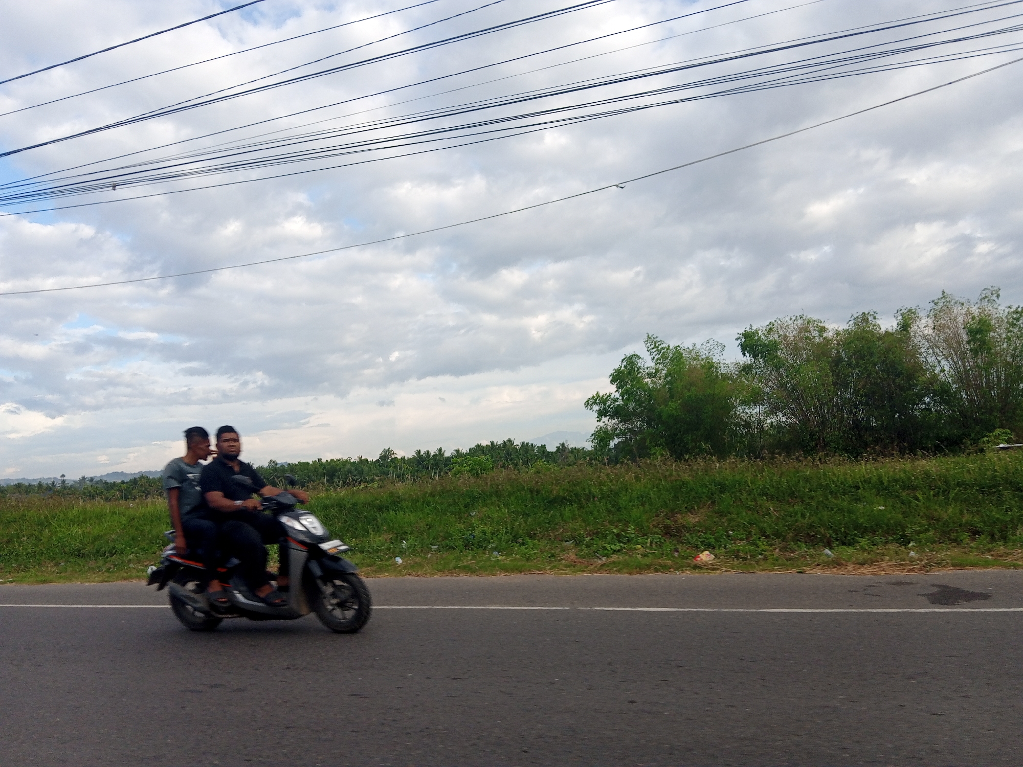 The image depicts two individuals riding a scooter on a road in what appears to be a rural area near Sagoe, Indonesia. The road surface is asphalt and has a white line marking the lane. A grassy verge separates the road from a field of vegetation, with trees and shrubs visible in the background. Overhead, power lines stretch across the sky, which is filled with clouds. The foreground of the road shows some dark patches. The scooter and its occupants are in motion.