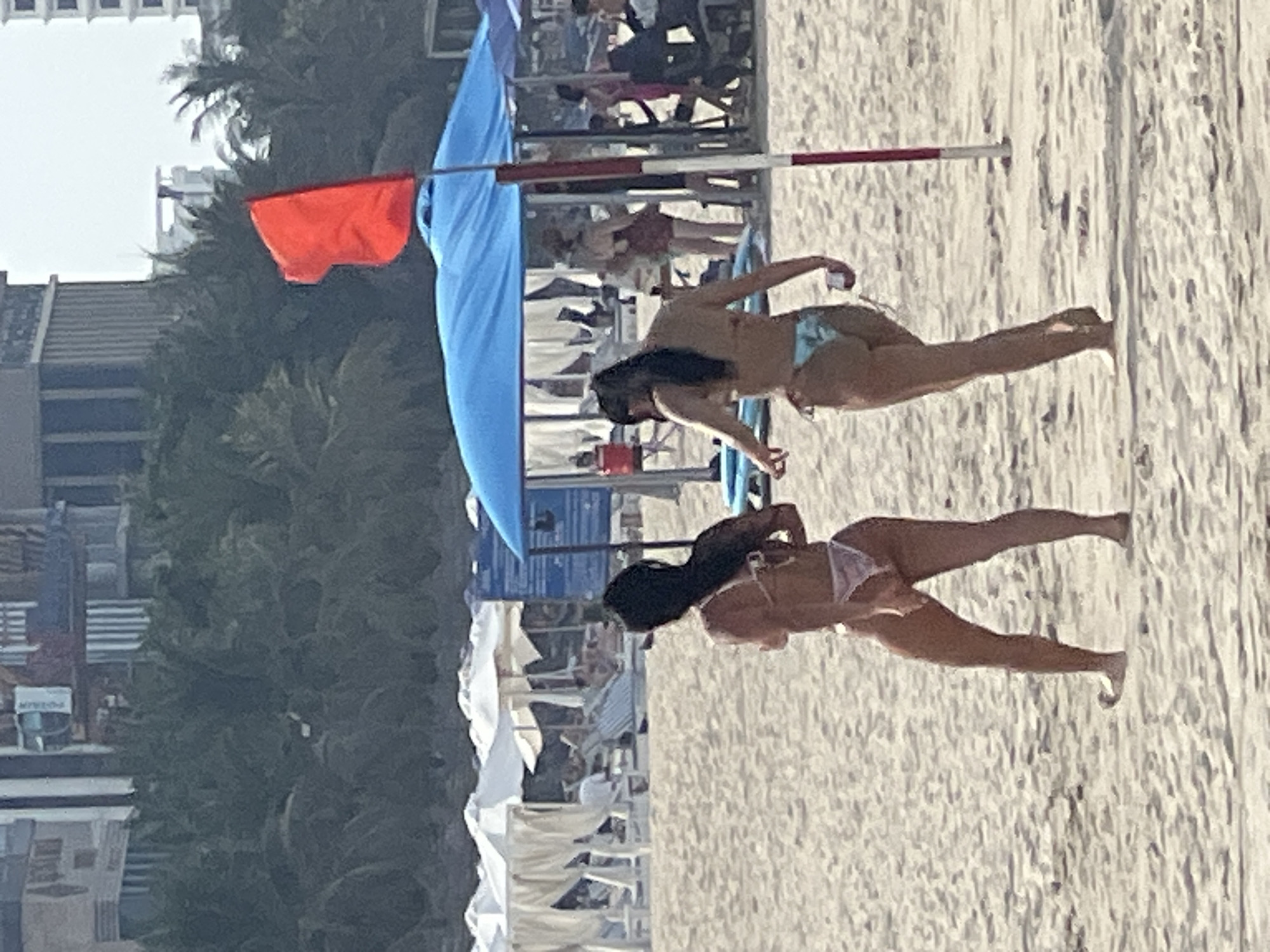 The image shows a beach scene in Da Nang, Vietnam. Two women are the primary subjects, both wearing bikinis. The woman in the foreground is in a white bikini and is on her hands and knees. The woman behind her is in a light blue bikini and is in a similar pose, with her legs extended. Both women have dark hair.
The setting is a sandy beach with the ocean not visible. In the background, there are beach chairs and umbrellas, suggesting a populated area. A blue canopy and a red flag on a pole are also visible, likely indicating a lifeguard station or a restricted swimming area. The overall impression is of a sunny day at the beach. The image is vertically oriented, and the resolution is somewhat low, giving it a slightly grainy appearance.