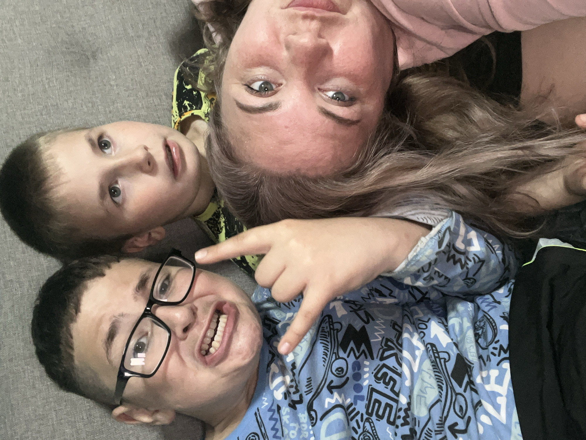 In this close-up, top-down selfie, three people are lying on a textured grey surface, likely a couch. Two boys are in the foreground, and a woman is positioned behind them, her head tilted upside down relative to theirs.
The boy in the bottom left is wearing glasses and a blue t-shirt with a black and white graphic pattern. He is making a "rock on" hand gesture with his right hand and has a wide, toothy grin. His eyes are looking directly at the camera.
To his right and slightly above, another boy is visible, looking up and to the left. He has light brown hair and appears to be smiling.
The woman at the top of the image has long, light brown hair and is wearing a pink top. Her eyes are open, and she is looking slightly off to the side. Her expression is somewhat neutral, perhaps a bit amused.
The overall impression is a casual, informal moment, likely a family selfie. The lighting suggests it's daytime, and there are no indicators of specific weather or an outdoor location. The focus is entirely on the faces and expressions of the individuals. No visible text is present in the image.