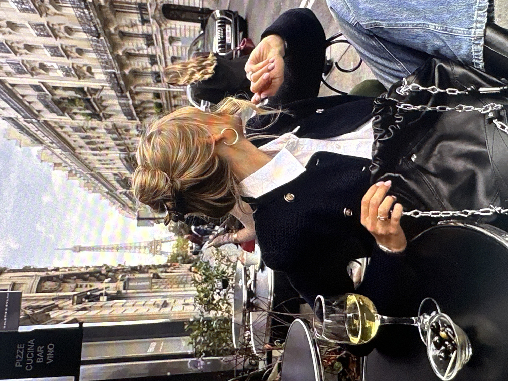 The image shows a scene at an outdoor cafe in Paris, France, with the Eiffel Tower visible in the background. A woman with blonde hair styled in a bun is seated at a table. She wears gold hoop earrings, a white collared shirt, and a black jacket with gold buttons. A black leather handbag with a silver chain strap sits on the table. Two wine glasses are visible on the table, one containing white wine and the other containing olives. Another woman, with long blonde hair, is partially visible in the background. The cafe is situated along a street lined with buildings. A sign with the words "PIZZE CUCINA BAR VINO" can be seen.