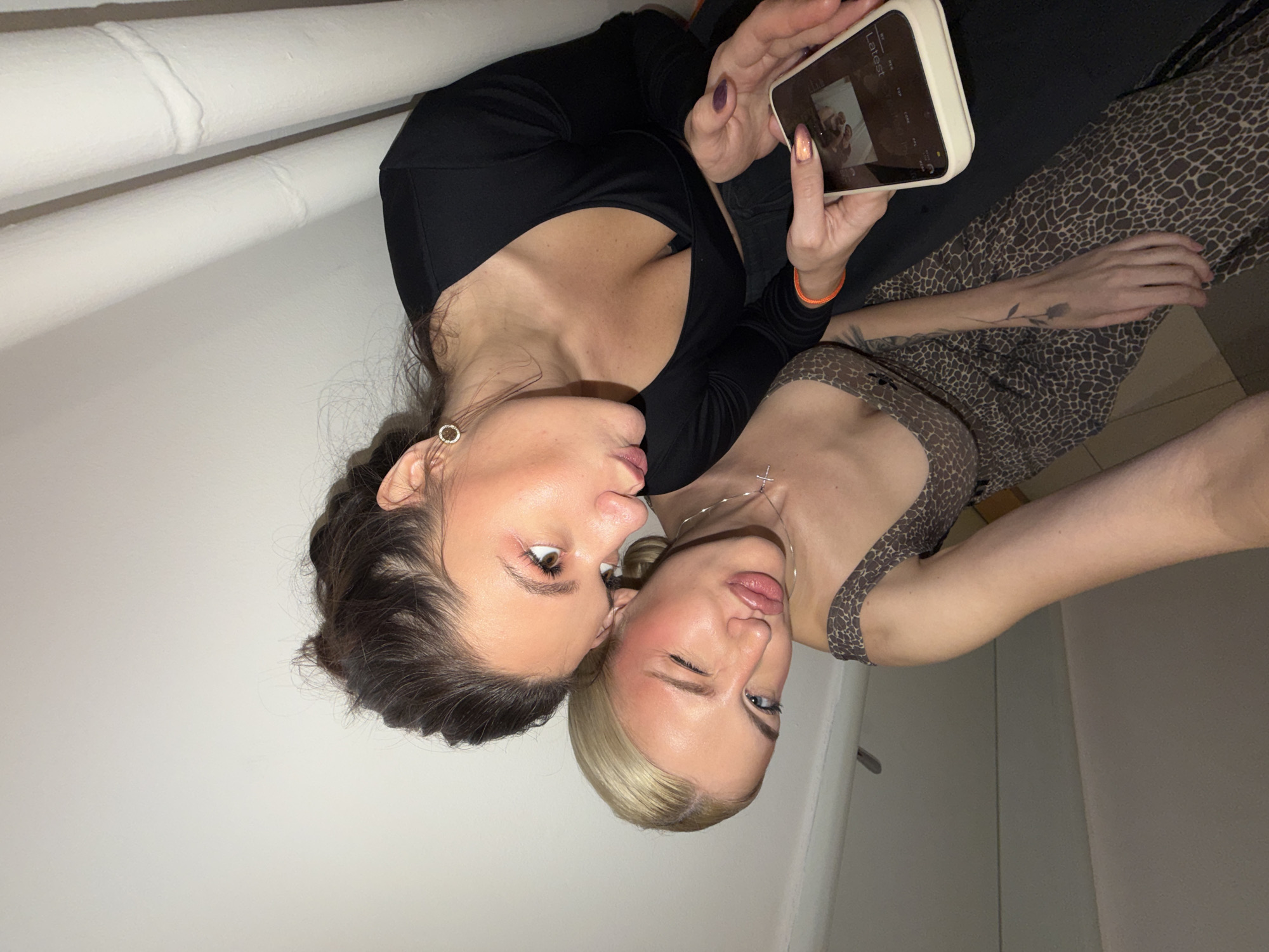 Two young women are captured in a playful selfie-style photograph indoors in Belgrade, Serbia. The image is tilted, suggesting a dynamic, casual pose.
The woman on the left, with dark brown hair styled in a braid, is wearing a black long-sleeved top and a round, hoop-style earring. She is making a "kissing" face with her lips pursed and eyes wide open. She holds a white-cased smartphone in her right hand, with her fingers visible. The phone screen displays the word "Latest" at the top left, indicating a gallery or camera interface. Her nails are painted a dark purple.
Leaning into her, the woman on the right has blonde hair and is wearing a sleeveless top with a brown and black animal print (likely leopard or giraffe). She has a thin silver necklace with a cross pendant. Her face is turned towards the camera, with one eye partially closed in a wink and her lips also playfully pursed. A tattoo of a rose is visible on her right forearm. Her nails are a shimmering light color.
In the background, two white pipes run horizontally across a white wall, contributing to a simple, somewhat utilitarian indoor setting. The lighting appears to be artificial, casting soft shadows. The overall mood is affectionate and friendly.