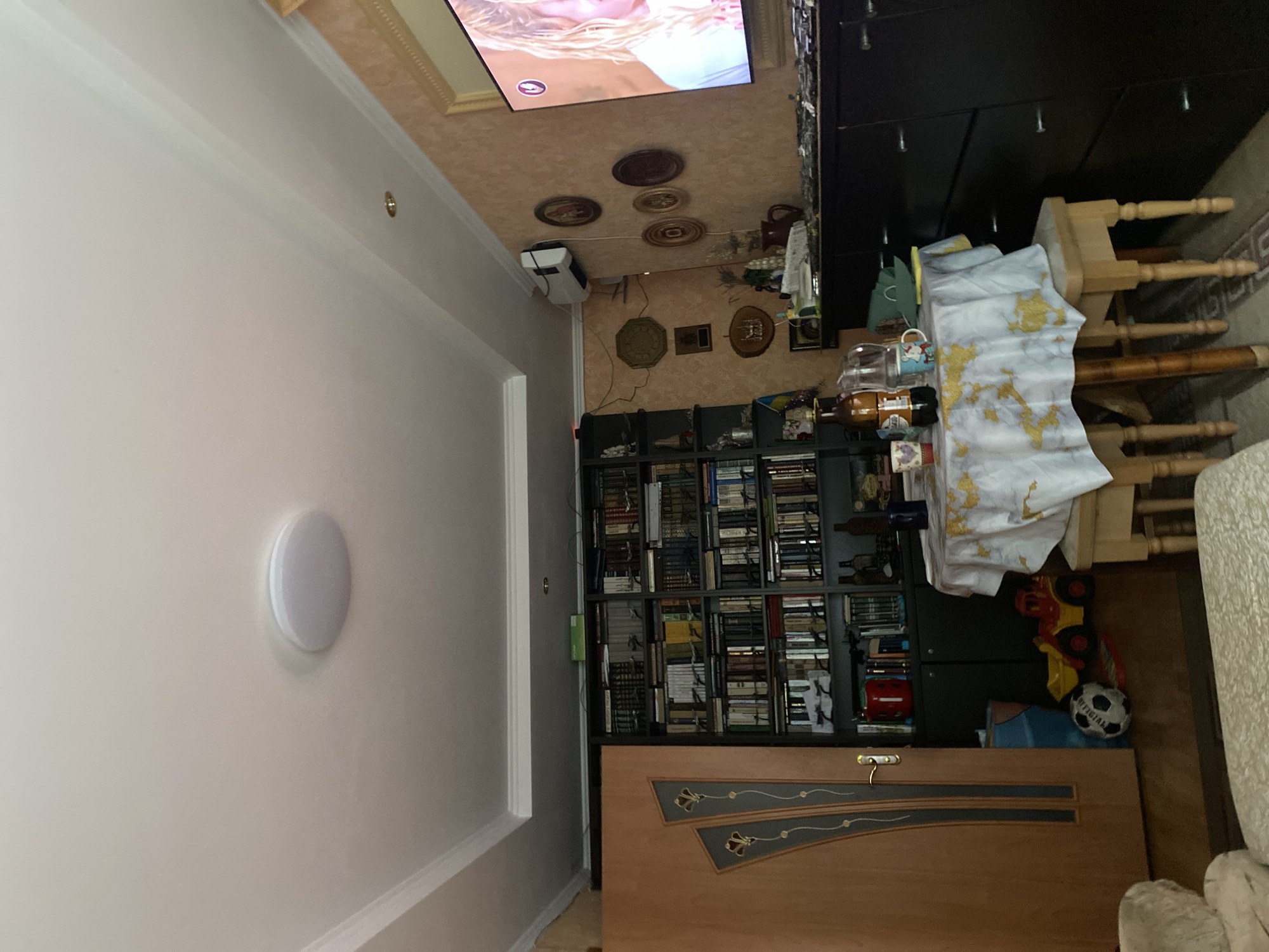 The image depicts an indoor scene, likely a living room, in a dwelling located in Unknown, Unknown. The room is furnished with a bookshelf filled with books, a television displaying an image of a woman, a small round table covered with a tablecloth and set with drinks and cups, and a dark dresser. A doorway with decorative panels is visible on the left side of the frame. Children's toys, including a soccer ball and a toy truck, are present on the floor. Decorative items, such as framed pictures and plates, adorn the wall. The ceiling features recessed lighting. The overall impression is of a lived-in space.