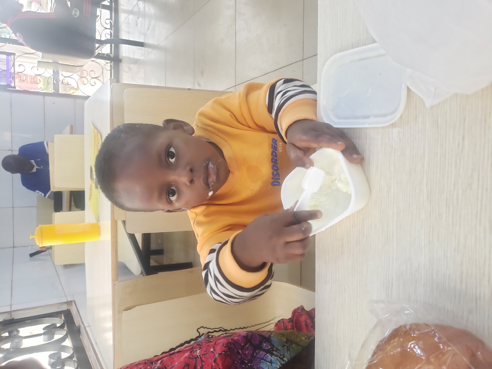 The image depicts a young Black child, likely a boy, wearing an orange sweatshirt with striped sleeves. He is seated at a light-colored table, with his face close to the camera, and appears to be eating or playing with a white food item from a white container using a small white spoon. Some of the food is visible on his lips and chin. To his left, partially visible, is a person wearing a colorful patterned garment. In the foreground, to the left, is a round bread item wrapped in clear plastic. To the right of the child, on the table, is another white container with its lid off. In the background, several other people are present, including a man in a blue shirt sitting at a table and another person partially visible on the right. The environment suggests an indoor setting, possibly a dining area or communal space, with light-colored tables and chairs, and a tiled floor. The overall impression is of a casual, everyday moment. The presence of the bread item and the communal dining setup could indicate this is a school or community center in Sabon Tasha, Nigeria.