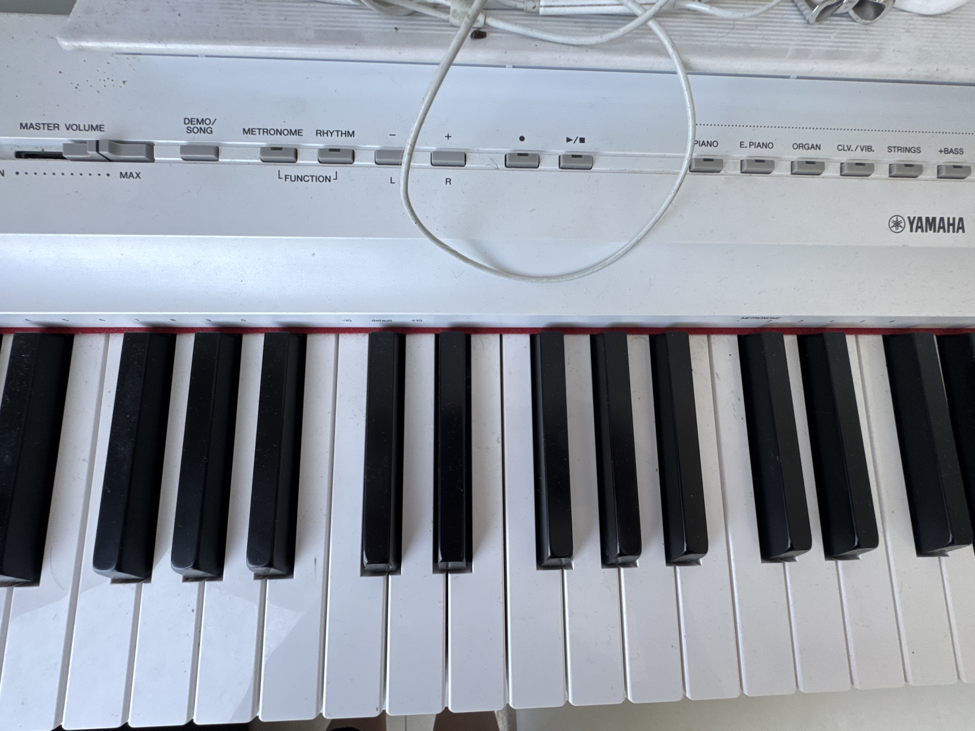 A close-up, high-angle, eye-level shot showcases the control panel and keys of a white Yamaha digital keyboard. The control panel features various buttons and sliders for functions such as Master Volume, Demo/Song, Metronome, Rhythm, and instrument selections like Piano, E. Piano, Organ, Clv./Vib., Strings, and Bass. A power cord is visible draped across the top of the keyboard. Below the control panel are the black and white keys of the instrument. The scene appears to be indoors, on a flat surface, with no other discernible objects or environmental details present. The keyboard is positioned on a surface, likely a table or stand.