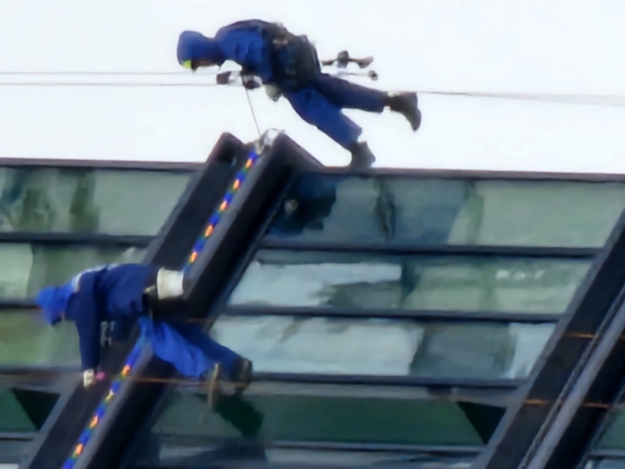 The image shows two individuals in blue protective suits with harnesses, engaged in window cleaning on the exterior of a modern building. The individuals are suspended by ropes and appear to be working at different levels. One person is higher up, leaning forward and seemingly in motion, while the other is positioned below and to the side, also in a dynamic pose. Both are equipped with safety gear, including helmets and harnesses.
The building's architecture features large glass panels and dark, angled structural elements. A string of colorful lights, possibly decorative or functional, is visible along one of the angled supports. The sky is overcast, suggesting a cloudy day. The overall scene depicts a high-risk operation, likely window washing or maintenance on a tall structure. The focus is on the workers and their activity, highlighting the challenges and safety measures involved in working at heights. No text is visible in the image. The location is specified as Moscow, Russia.