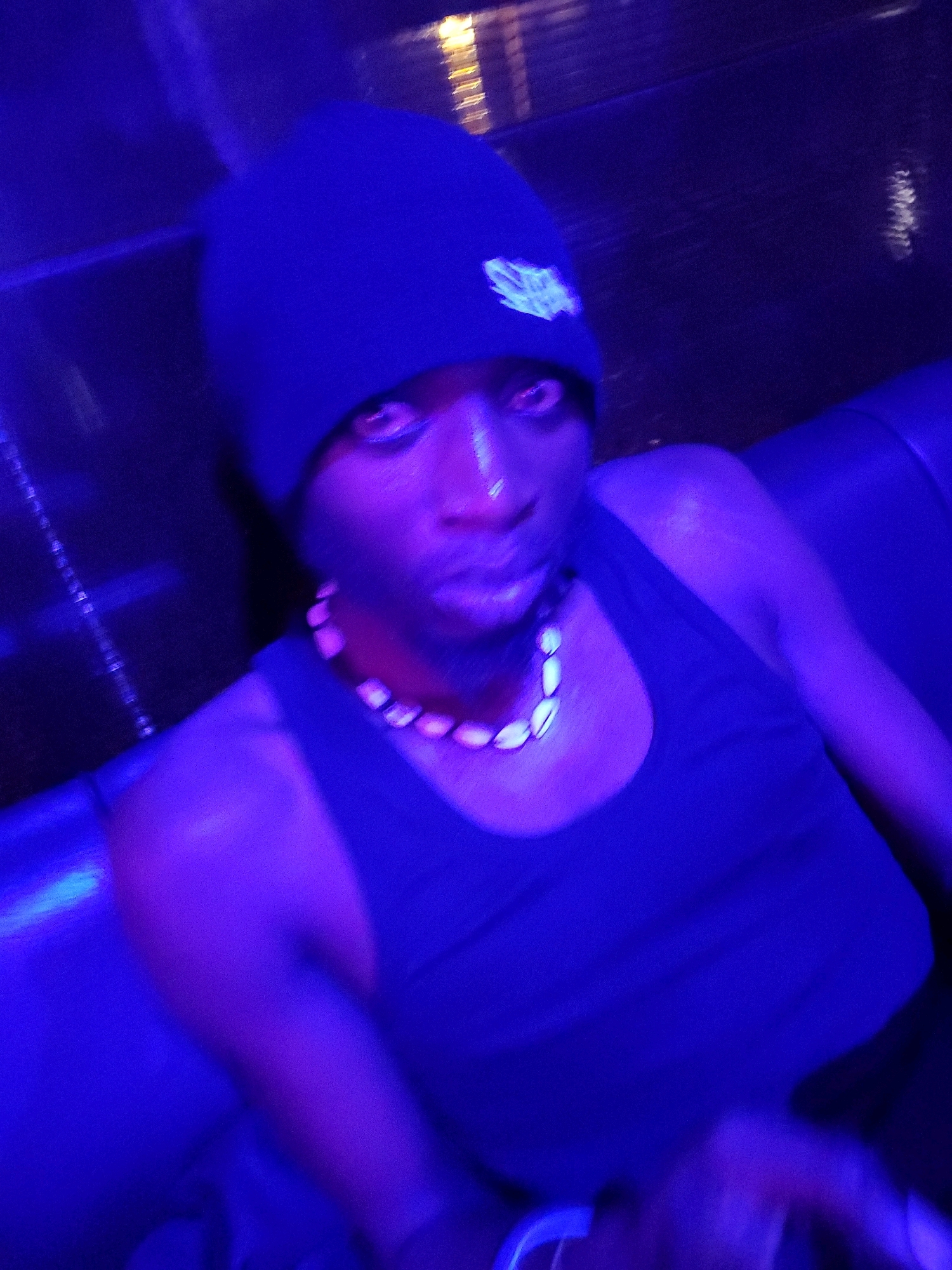 A low-angle, slightly blurry, medium shot captures a young Black male from the chest up, wearing a dark blue beanie with a white emblem, a dark blue tank top, and a shell necklace, seated on a dark blue couch. The scene is dimly lit with strong blue and purple ambient lighting, suggesting a nightclub or similar venue. The man is looking directly at the camera with his mouth slightly open. The background consists of a dark, textured wall and the back of the couch. There are no other individuals visible. The specific location in Jos, Nigeria cannot be determined from the visual information provided.
