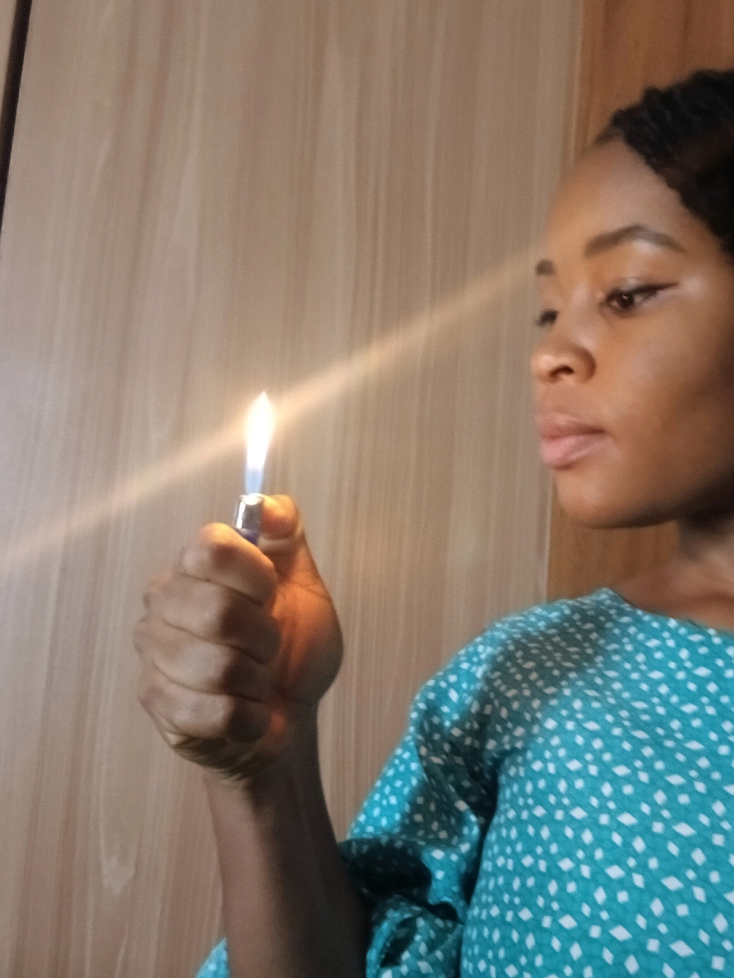 A medium shot captures a woman holding a lit lighter in her right hand, the flame casting a bright glow. The woman's face is turned slightly away from the camera, her gaze directed off-frame to the right. She has dark hair styled in braids and is wearing a teal dress with a white polka dot pattern. The background consists of a wooden wall, suggesting an indoor setting in the city of Gura Top, Nigeria.