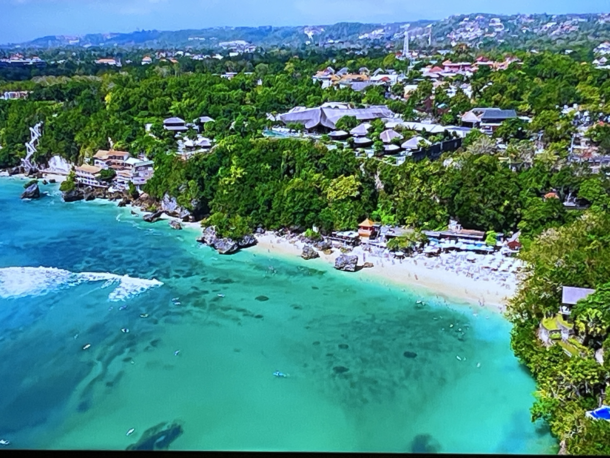 This is an aerial drone shot of a tropical coastline. The turquoise water is clear and shallow, revealing darker patches of coral or rocks below. Small boats are scattered across the water, and waves gently break near the shore. A sandy beach is visible, lined with numerous beach chairs and umbrellas. Buildings, some with distinctive thatched roofs, are nestled amongst lush green trees that cover the hillsides. The architecture suggests a resort or hotel complex. The scene appears to be during the day, with clear, bright lighting indicating fair weather. The overall impression is one of a tranquil and idyllic holiday destination. There is no visible text in the image.