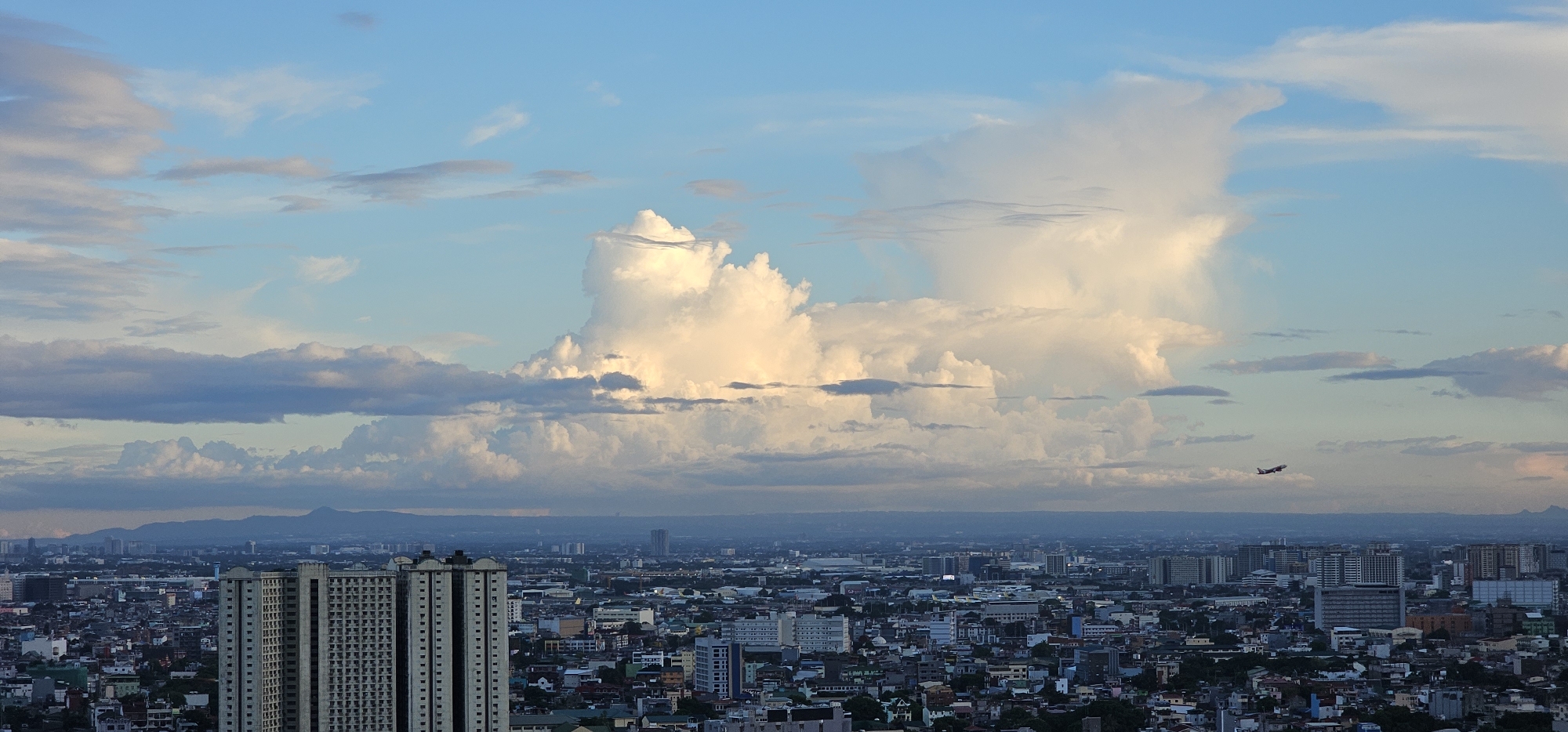 A panoramic cityscape of Makati, Philippines, is captured under a vast expanse of sky. Dominating the upper half of the frame are dramatic clouds, ranging from wispy stratocumulus to towering cumulonimbus formations illuminated by warm, late-afternoon light. The sky itself transitions from a pale blue at the zenith to a softer, warmer hue near the horizon, indicative of an approaching sunset.
The city below is a dense tapestry of buildings. In the foreground, two imposing, rectangular high-rise residential buildings stand out, their facades textured with repetitive window patterns. These structures are flanked by a sprawling urban landscape characterized by a mixture of lower-rise residential homes and commercial buildings, creating a dense and varied architectural texture. The city extends towards a hazy horizon, where a range of distant mountains can be faintly discerned.
Adding a dynamic element to the scene, a commercial airplane is visible in the mid-right portion of the image, appearing as a small silhouette against the sky and clouds, indicating it is either taking off or landing. The overall atmosphere is one of urban expanse meeting the grandeur of nature, with the city bathed in the soft, diffused light of the sky.