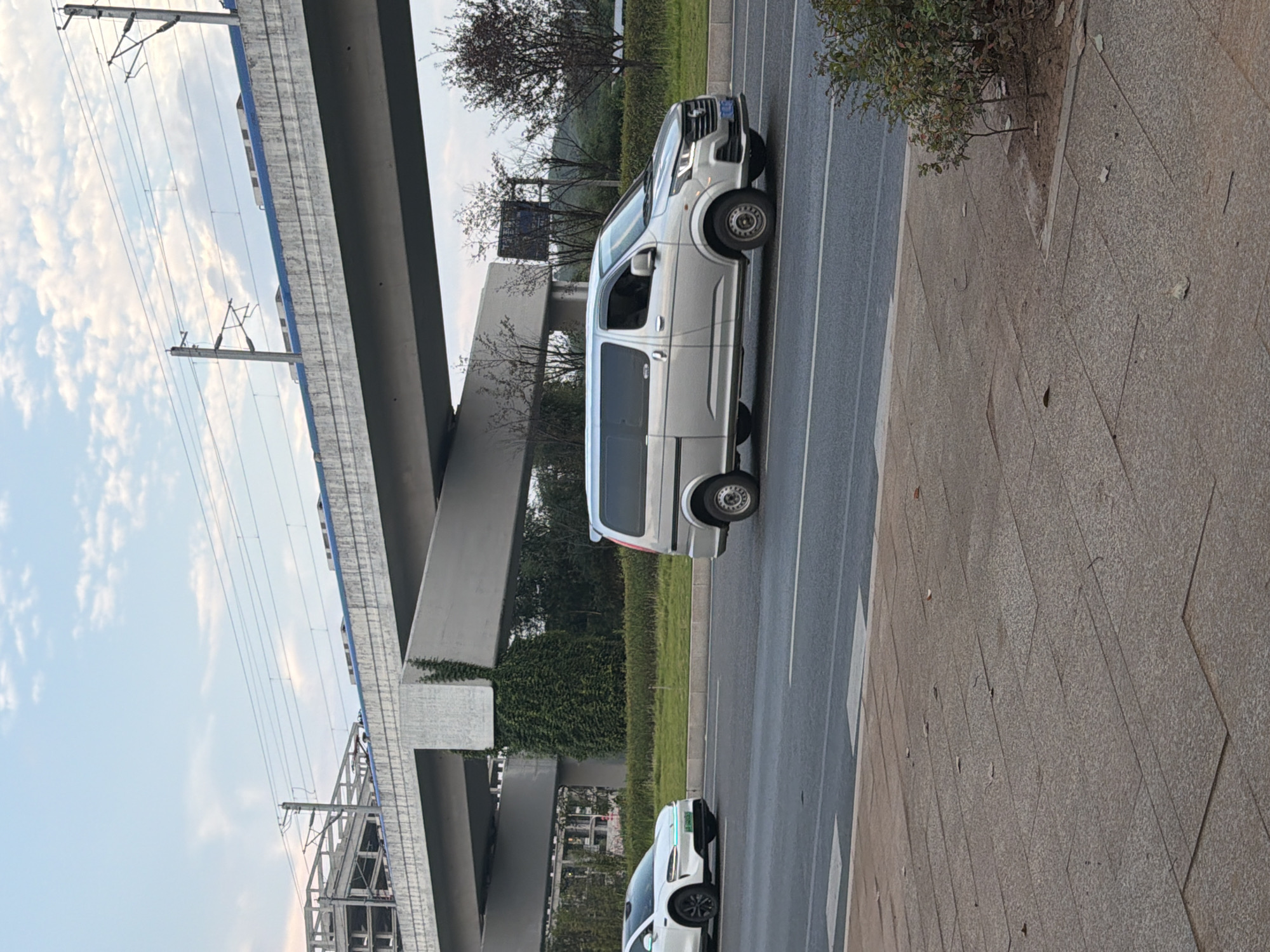 In the city of Jiangjin, China, a white electric vehicle is parked on the left side of a road, facing right. Adjacent to it, a silver minivan is driving on the same road, also facing right. The road is flanked by a sidewalk and a median strip with green grass and trees. An elevated concrete structure, likely a bridge or overpass, spans across the background, with train tracks and overhead power lines visible on top. The sky is partly cloudy.