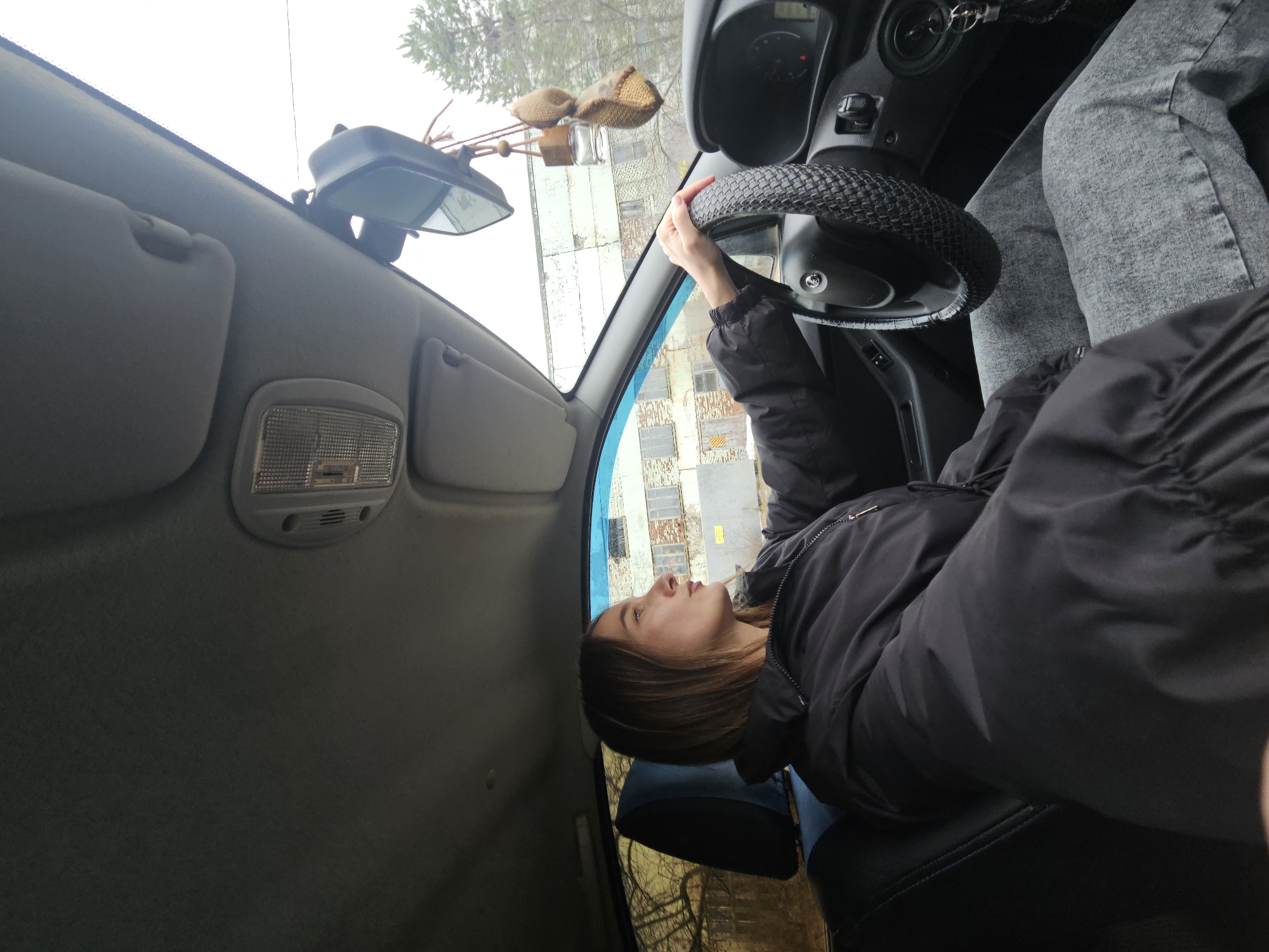The image captures a young woman seated in the driver's seat of a car, viewed from a slightly elevated, side-on perspective within the vehicle. The woman, with shoulder-length brown hair, wears a black puffer jacket and light grey jeans. Her left hand rests on the steering wheel, which is covered with a black, textured material. She is looking upwards and to her right, seemingly lost in thought or observing something outside the frame, with a calm, somewhat pensive expression.
The car's interior details are visible, including the dashboard with gauges, a rearview mirror from which a woven, light-brown decorative item with a small glass bottle hangs, and sun visors. An overhead interior light fixture is also present. The car seats are dark, possibly black.
Through the windshield, an overcast sky dominates the upper portion of the view, with some bare tree branches visible. A multi-story building with a light-colored, possibly industrial or residential, weathered facade and numerous windows is seen in the background. The overcast conditions suggest a cool, daytime setting without direct sunlight. There is no visible text in the image. The scene is set in Chaikovsky, Russia.