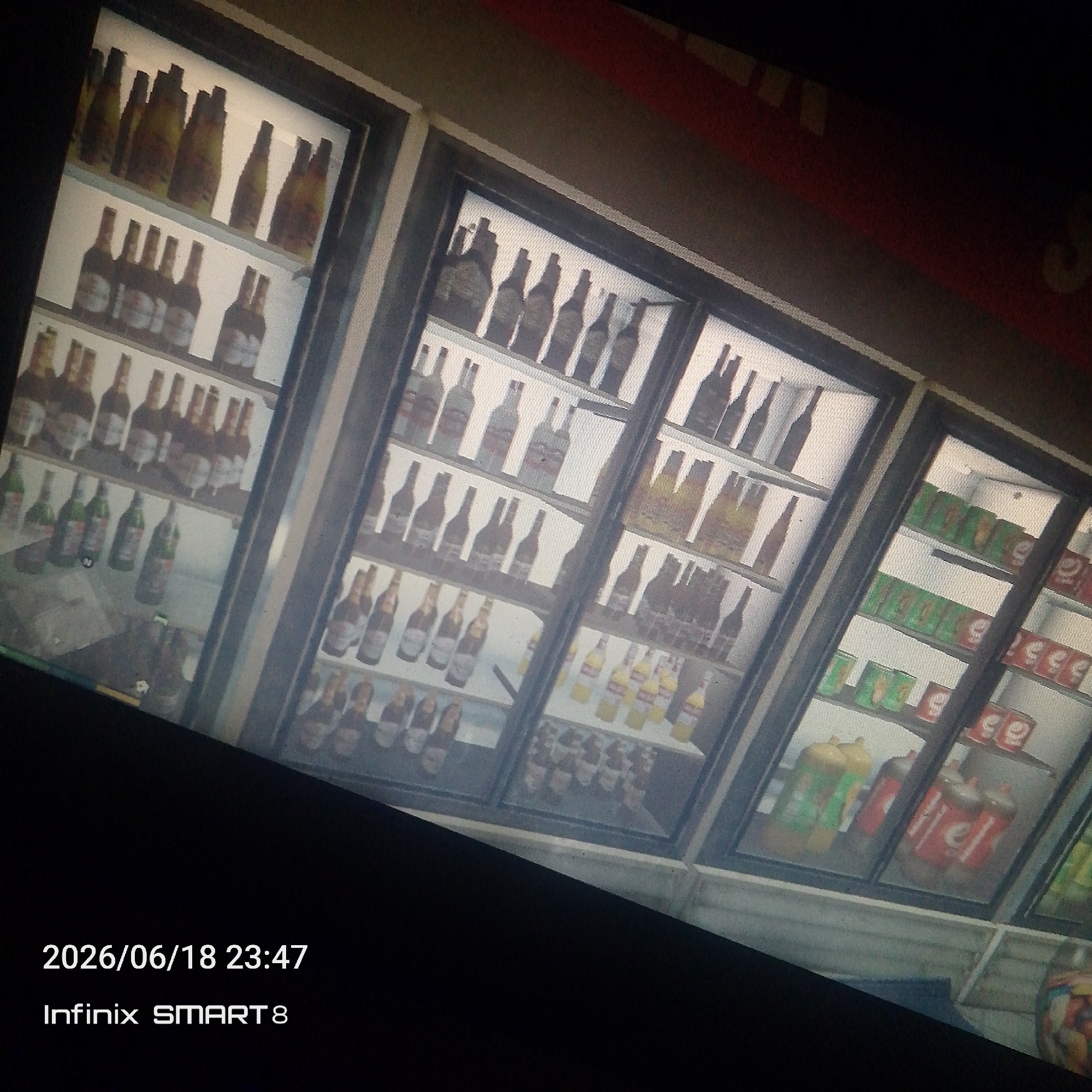 This image is a photograph of a refrigerated display case filled with various beverages, likely in a store or bar. The display case is divided into multiple shelves, each stocked with bottles and cans. On the top shelves, there are rows of glass bottles, some with yellow labels and others with darker labels. The middle shelves contain more bottles, some appearing to be beer bottles, and further down, there are rows of green and red cans, suggestive of soft drinks or energy drinks. On the lower shelves, larger bottles, possibly of juice or soda, are visible, along with more cans. The lighting inside the display case illuminates the products. The overall scene is viewed at an angle, with part of the surrounding environment dark and out of focus. In the lower left corner of the image, a timestamp "2026/06/18 23:47" and device information "Infinix SMART 8" are superimposed. The image does not contain any information that would indicate the specific location in the city of Jos, Nigeria.