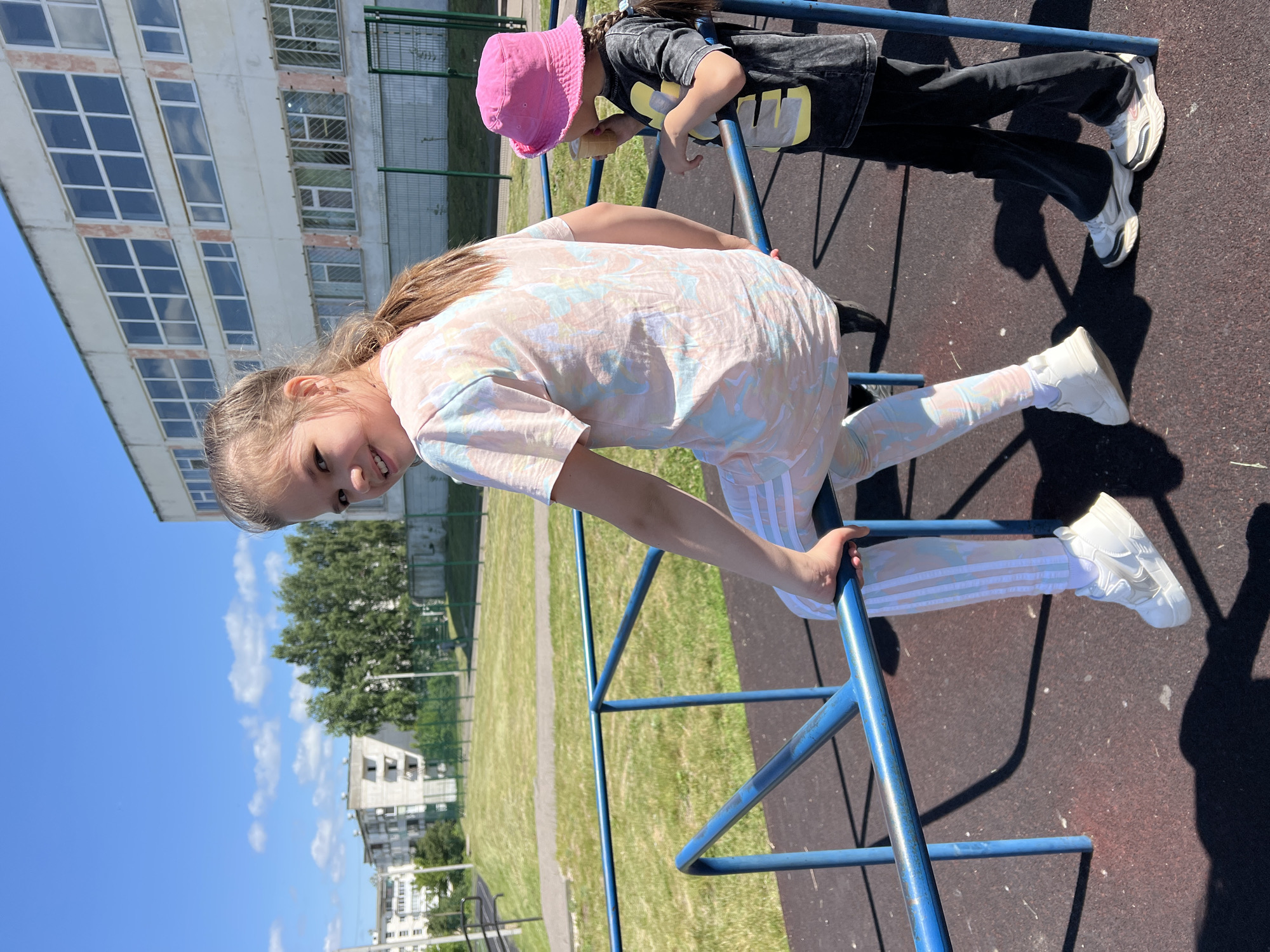 Two young girls are on a playground in Chaikovsky, Russia. The girl in the foreground, on the left, is sitting on a blue metal bar and looking over her shoulder at the camera. She has blonde hair in a ponytail, is wearing a pastel tie-dye t-shirt and matching leggings, and white sneakers. To her right, a younger girl in a pink bucket hat and a dark t-shirt is standing and eating an ice cream cone. Behind them is a school building and a grassy area with a fence.