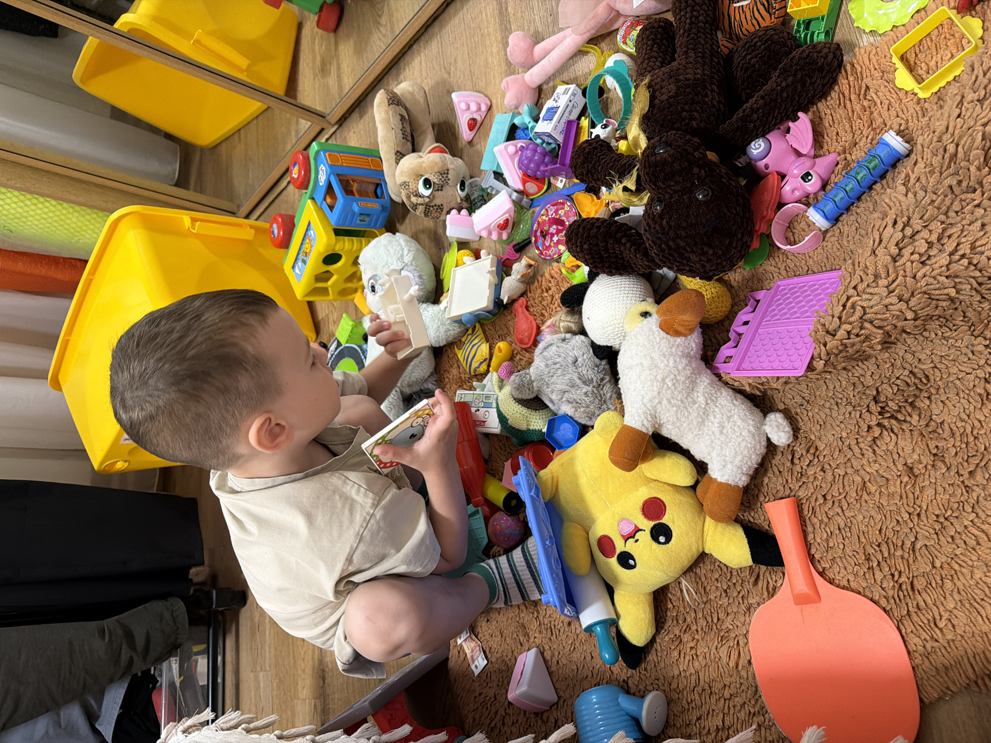A young boy is playing with a variety of toys on a fluffy brown rug. He is wearing a light-colored t-shirt and green and white striped socks, and is crouching down, engaged in examining a small white toy in his hands. Surrounding him are numerous colorful toys, including stuffed animals like Pikachu and a lamb, building blocks, toy food, and other miscellaneous playthings. The scene appears to be indoors, possibly in a child's room or play area, with a wooden floor visible beneath the rug and reflections of yellow storage bins in a mirror in the background. The lighting suggests it is daytime. The boy's focused expression and the scattered toys indicate an active play session.