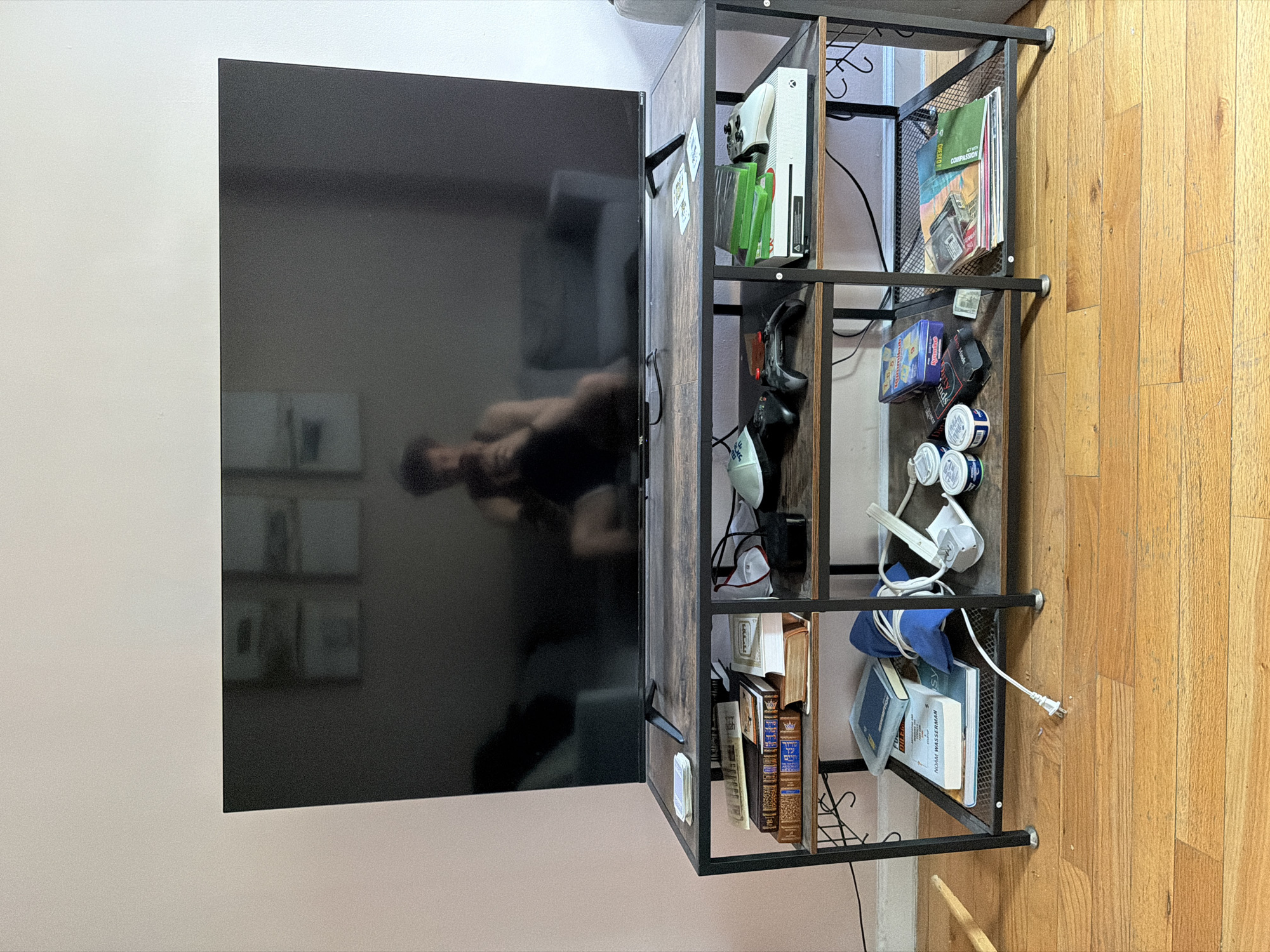 An indoor scene, likely a living space or bedroom in New York, United States of America, displays a large, dark TCL television screen mounted on a light-colored wall. A reflection of a person, holding a device to take the photograph, is visible on the television screen. To the right of the TV, a multi-tiered shelving unit, constructed from dark metal and faux wood panels, serves as a media console and storage.
The top shelf of the unit holds a white Xbox console and several green-spined Xbox game cases. Below this, a shelf contains two video game controllers (one black, one white with blue grips), a pair of glasses, headphones, and a white power strip. Further down, another shelf displays a blue cloth or bag along with cables, a power strip, and an electrical plug. Various small boxes, including one partially labeled "Cards Against Humanity," and three stacked cans (possibly tobacco products), are also present on the shelves. A dollar bill is tucked into the shelving. The bottom shelf is filled with books, some featuring distinct Hebrew text on their spines, and one clearly showing the author "NOAM WASSERMAN." A magazine or book on an upper shelf is titled "ACT WITH COMPASSION." The floor beneath the shelving unit is made of light-colored hardwood.