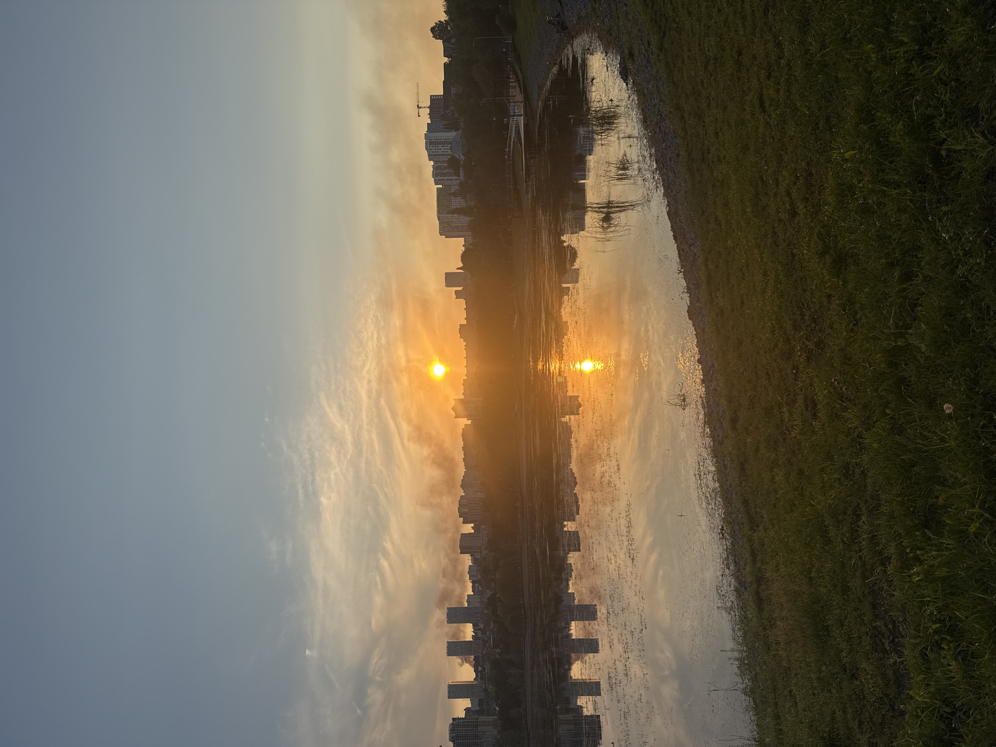 The image depicts a city skyline reflected in a body of water at sunset. The sun is low on the horizon, casting a warm orange glow across the sky and reflecting on the water's surface. A line of apartment buildings and other structures forms the city's silhouette against the colorful sky. Wisps of smoke are visible rising from behind the buildings, obscuring some of the cityscape. The foreground features a grassy embankment leading down to the water's edge, with some vegetation growing in the shallows. The water itself is calm, with ripples reflecting the light and the sky. This scene is likely from the city of Ufa, Russia, given the visual cues of urban development and natural landscape typical of the region.