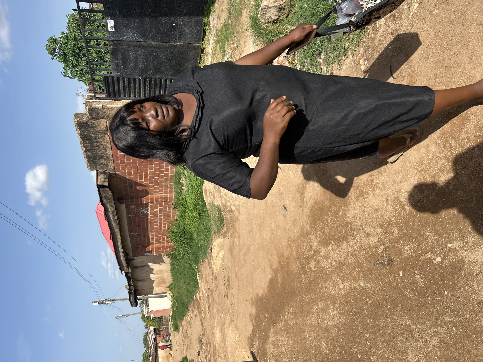 In Jos, Nigeria, a Black woman in a black dress walks on a dirt road. She has dark hair and is carrying a black bag in her right hand. The road is lined with red brick buildings, some with corrugated metal roofs. Green grass grows along the side of the road and a black metal gate is visible in the background. The sky is blue with scattered clouds. The overall scene depicts a street in a residential area.