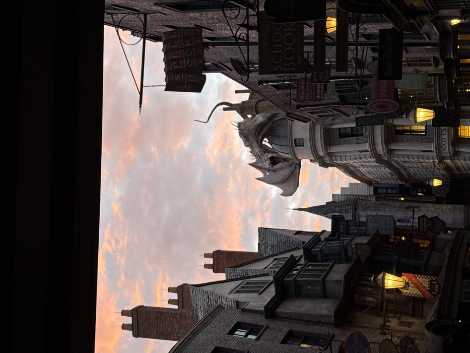The image displays a meticulously themed street scene, identified as Diagon Alley within Universal Studios Florida in Orlando, United States of America. The sky above exhibits a transitional lighting condition, characterized by pastel shades of orange, pink, and light blue, with visible cloud formations, indicative of either dawn or dusk.
A large, light-colored animatronic dragon is centrally positioned, perched atop a prominent domed structure identified as "Gringotts." The dragon's mouth is open, and its wings are partially spread. The street is flanked by multi-story buildings, constructed from brick and stone, featuring intricate architectural details such as gabled roofs, numerous chimneys, and bay windows.
Multiple ornamental street lanterns emit warm yellow light, illuminating the building facades and numerous hanging shop signs. Specific visible signs include "SPELL BOOKS" and "HOGWARTS SCHOOL BOOK" on the right side of the street. The top portion of the frame is obscured by a dark, horizontal structural element. No human figures are present within the visible area.