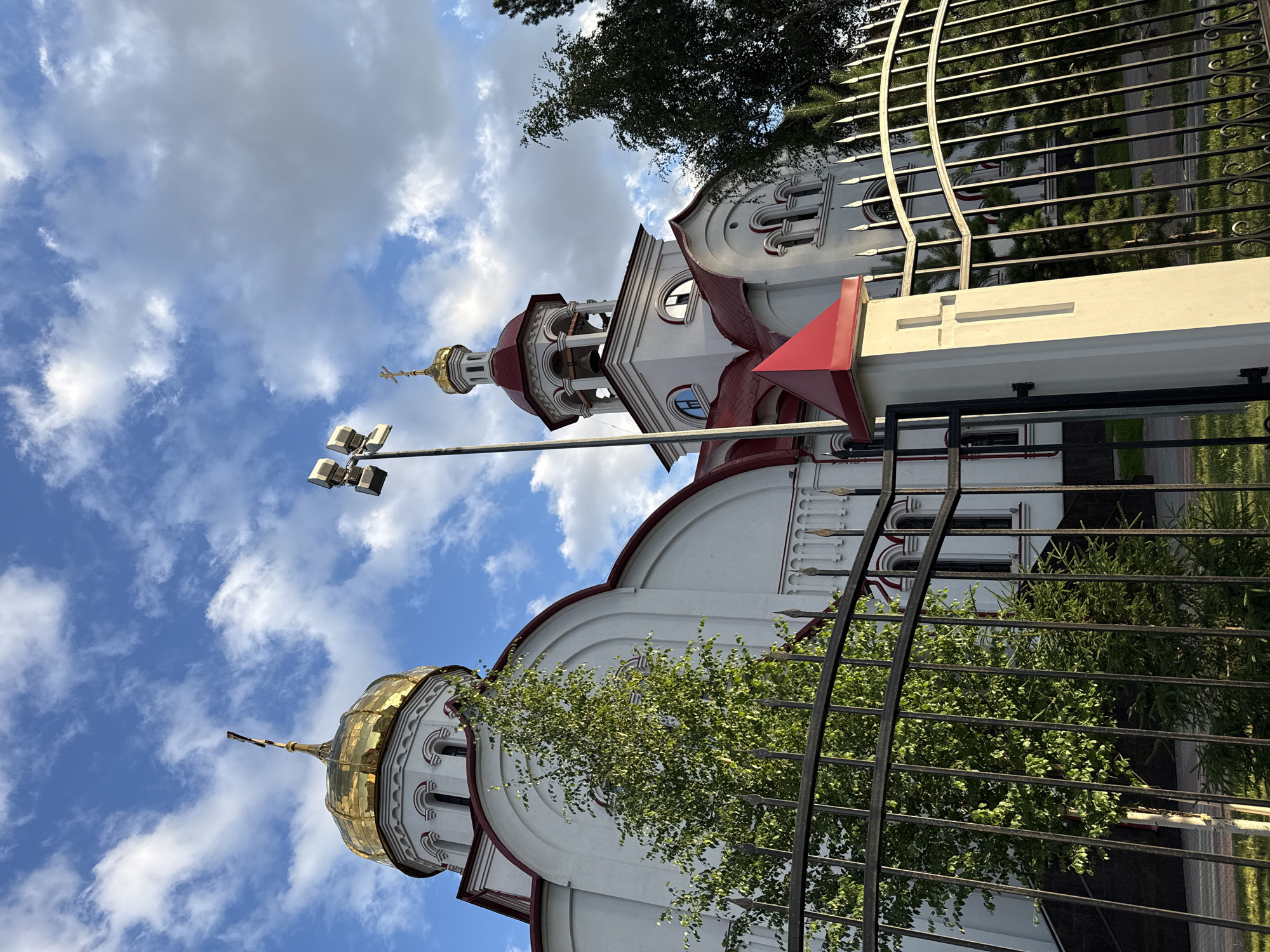 The image shows the Transfiguration Cathedral in Surgut, Russia, under a bright, partly cloudy sky. The cathedral is a white building with red roofs and golden domes. A black metal fence is in the foreground, with green trees behind it. The fence has decorative finials at the top and a white pillar with a cross motif. The sky is blue with scattered white clouds. The light suggests it is either morning or late afternoon, casting soft shadows. There are no people visible in the image.