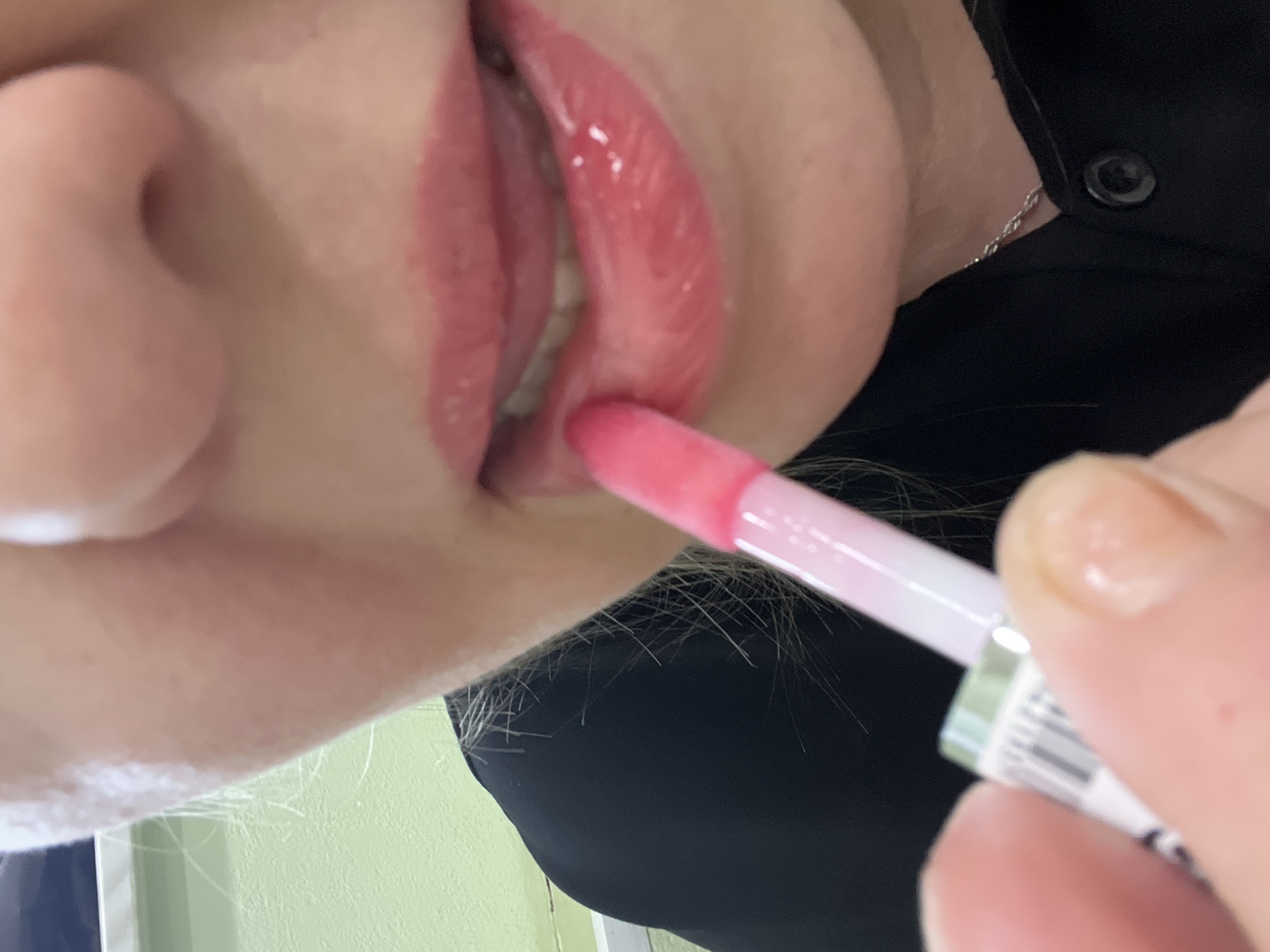 A close-up, high-angle shot captures a person in Chaikovsky, Russia, in the process of applying pink lip gloss. The image focuses on the lower part of the person's face, including their lips, nose, and chin, seen from a slightly upward perspective. The lips are already covered in a shiny, bright pink gloss, and the applicator, also pink and white, is positioned at the lower lip, suggesting the application is ongoing.
The person's skin tone is light, and strands of light-colored hair are visible near the chin and neck area. They are wearing a black garment, likely a top or dress, and a delicate silver chain necklace is visible around their neck. One hand, with neatly trimmed nails, holds the lip gloss bottle. A barcode is clearly visible on the side of the white and green lip gloss container, along with some indistinct small text.
The background appears to be a soft, light green surface, possibly an indoor wall, indicating an interior setting. The lighting is bright and even, casting minimal shadows and highlighting the glossy texture of the lips. The overall scene conveys a moment of personal grooming or makeup application.